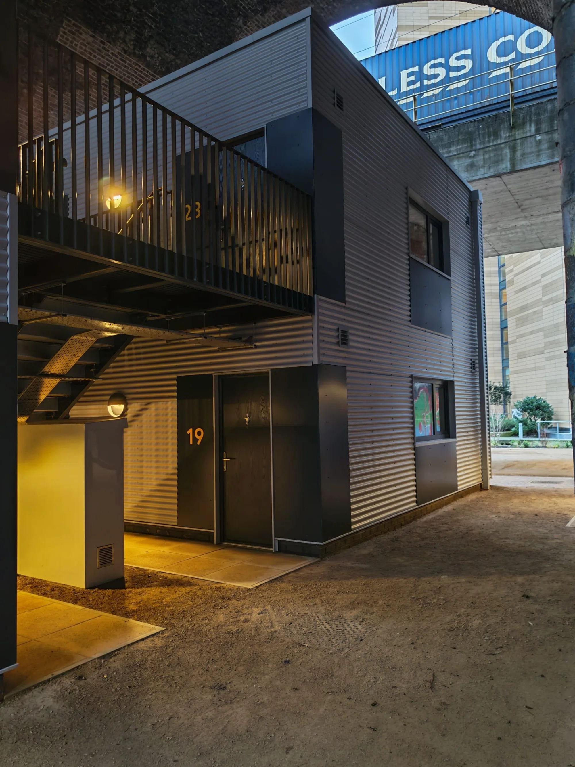 Two‑storey metal‑clad building under an archway, with doors labeled 19 and 23, an exterior staircase, balcony railings, and lit wall lamps. A large blue sign with partial text is visible above, along with nearby structures and walkways.