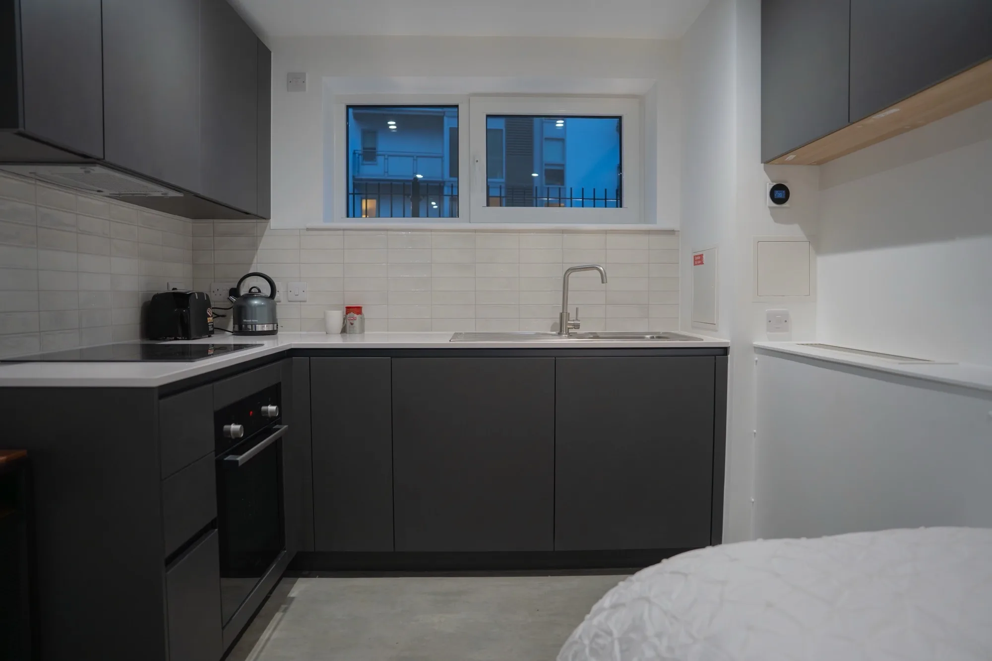 Compact kitchen with dark cabinets, an oven, stovetop, kettle, sink, tiled backsplash, and a window showing an exterior building. Light switches and a countertop appliance are visible on the walls and surfaces.