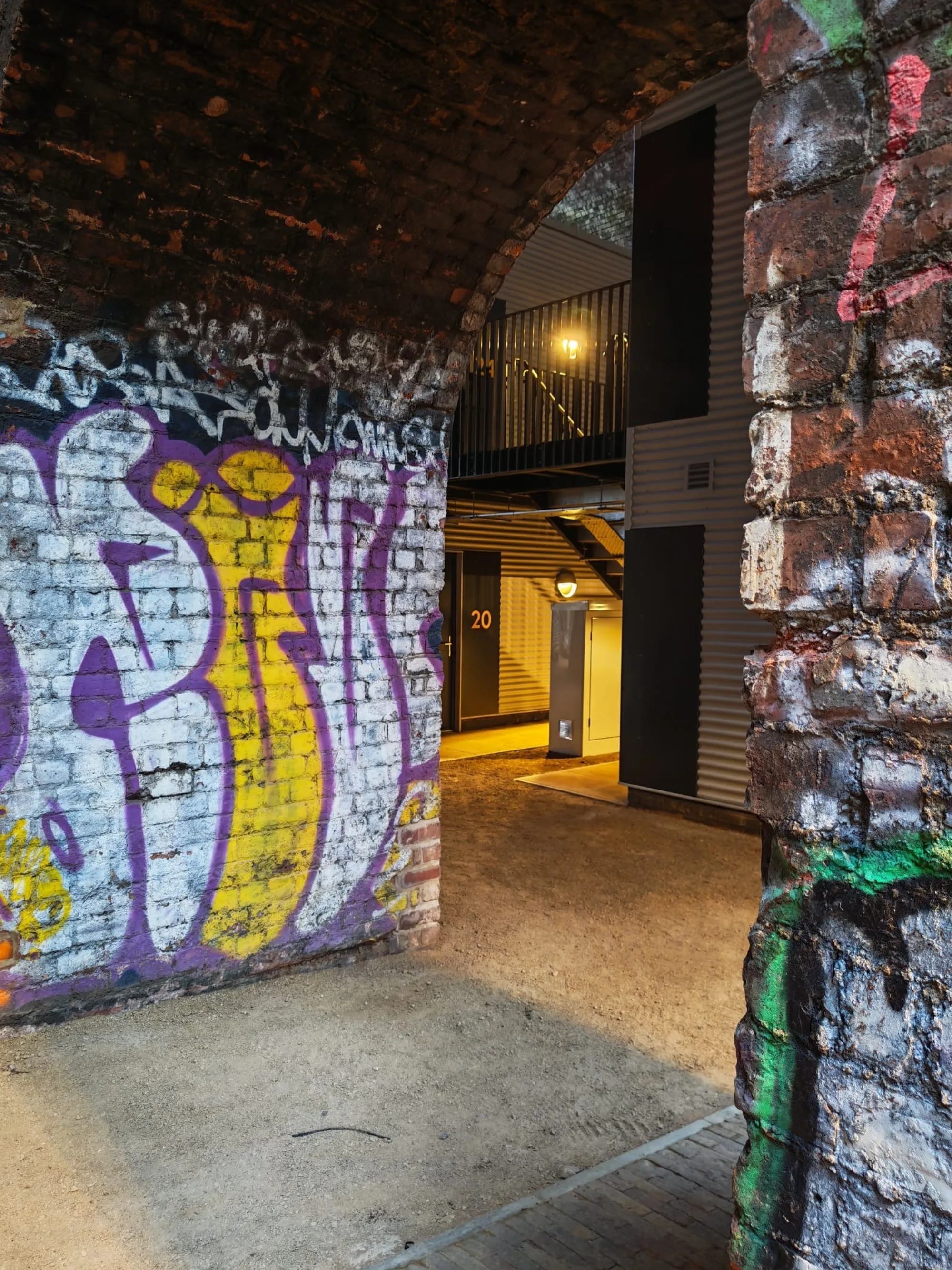 Brick archway with colorful graffiti in purple, yellow, white, and green framing a view of a metal‑clad building. A lit doorway labeled ‘20,’ an exterior staircase, upper walkway, and wall lights are visible inside the passage.