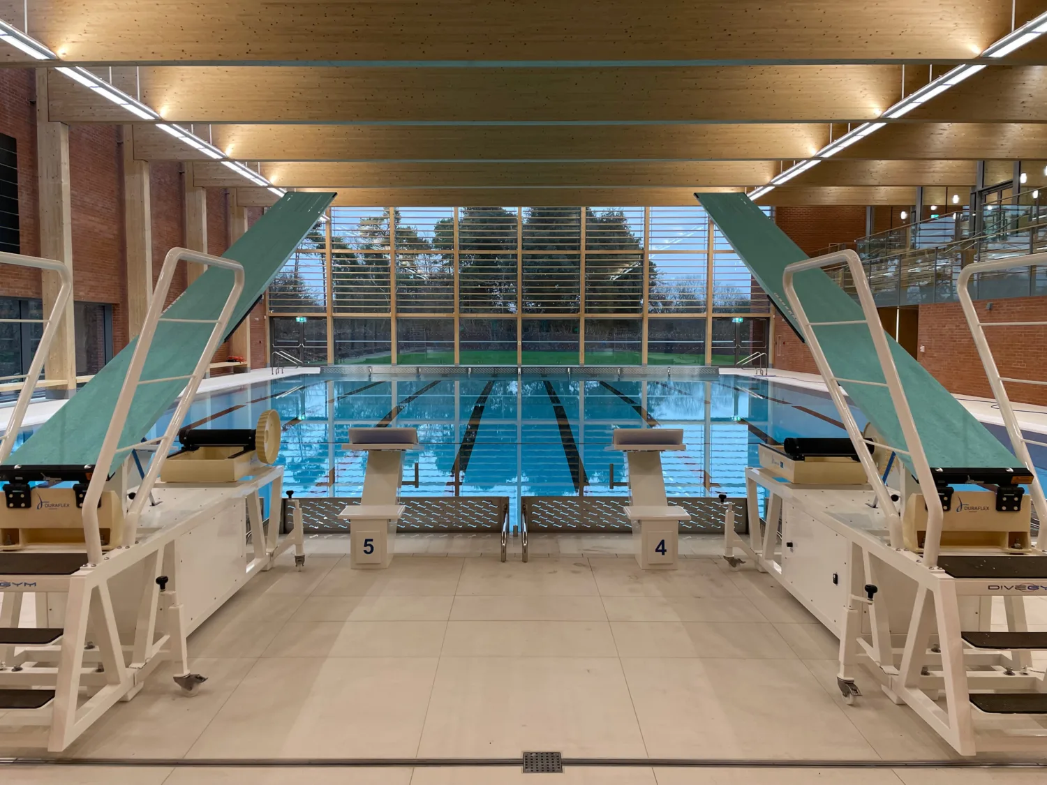 Swimming pool with diving boards, wooden roof overhead, full-length windows at end of pool