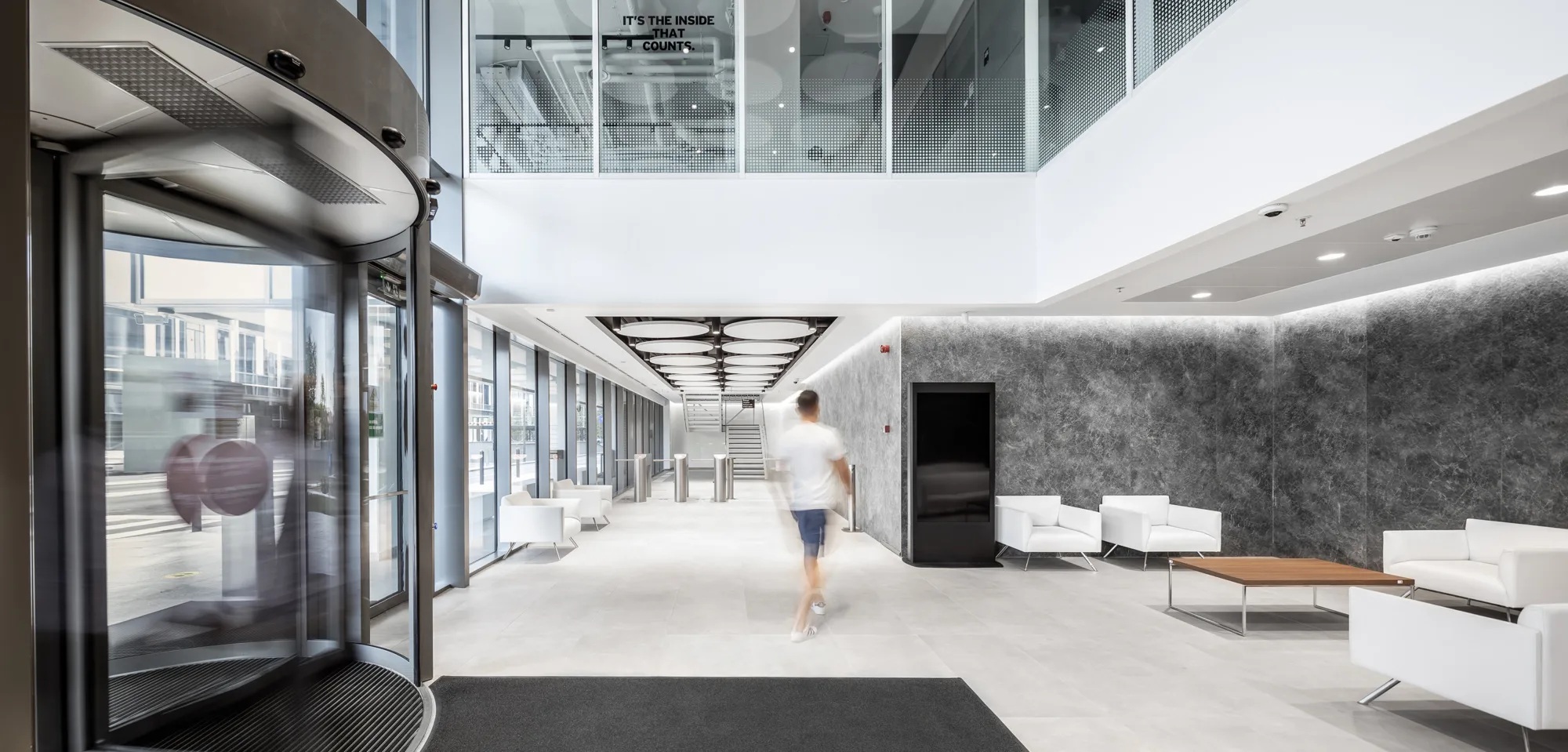 Inside the buildings lobby with white square seats and sofas, grey walls, white floors and security gates in the background