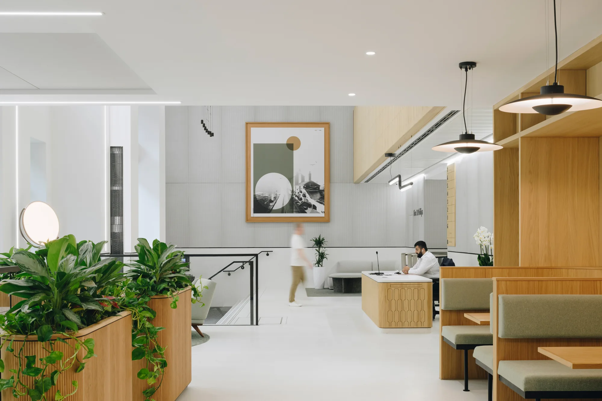 Modern office space with wooden furniture, green plants, and white walls. A person walks past a staircase leading down. Another person sits at a desk on the right. A large framed artwork is on the back wall.