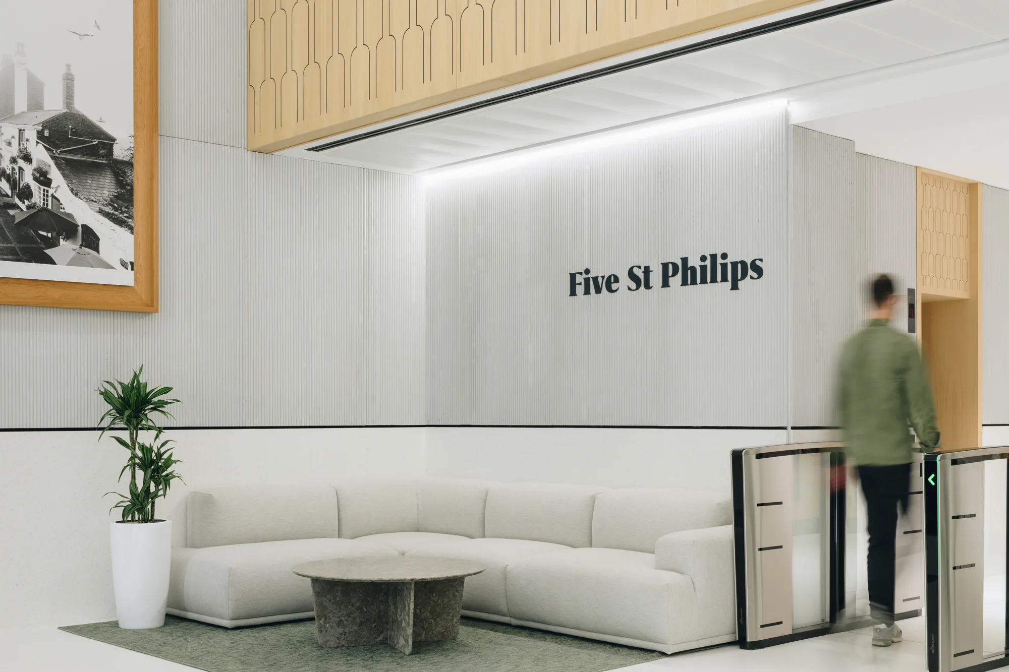 Modern office lobby with a white sectional sofa, round stone coffee table, and potted plant on the left. Wall behind the sofa displays the text "Five St Philips." A person walks through an access gate on the right.