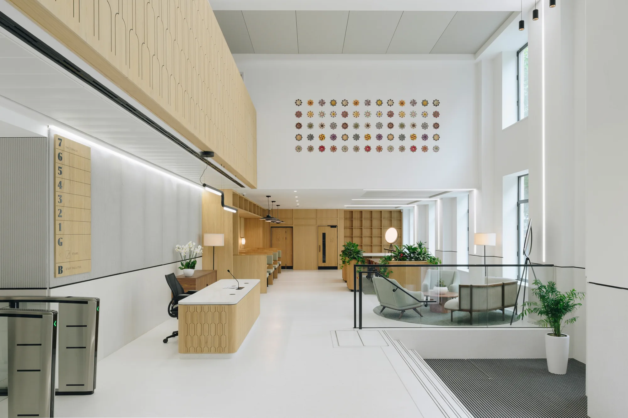 Modern office lobby with high ceiling and large windows. Left side has a reception desk, chair, computer, and turnstiles. Back wall has wooden cabinets and plants. Right side features seating, tables, lamps, and a wall with colourful circular patterns.