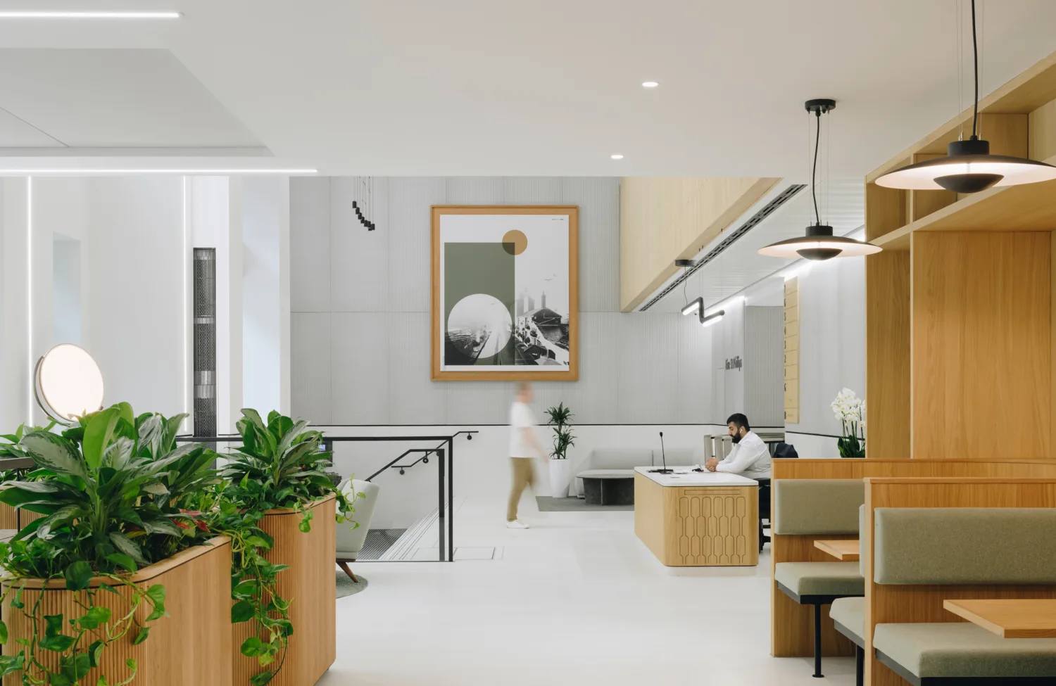 Modern office space with wooden furniture, green plants, and white walls. A person walks past a staircase leading down. Another person sits at a desk on the right. A large framed artwork is on the back wall.