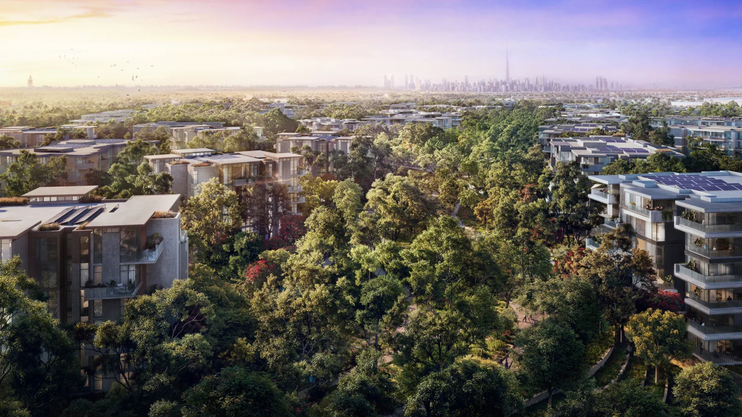 A wide aerial view of modern apartment buildings with flat roofs and some solar panels, surrounded by dense trees and walking paths, with a distant city skyline under a gradient sky.