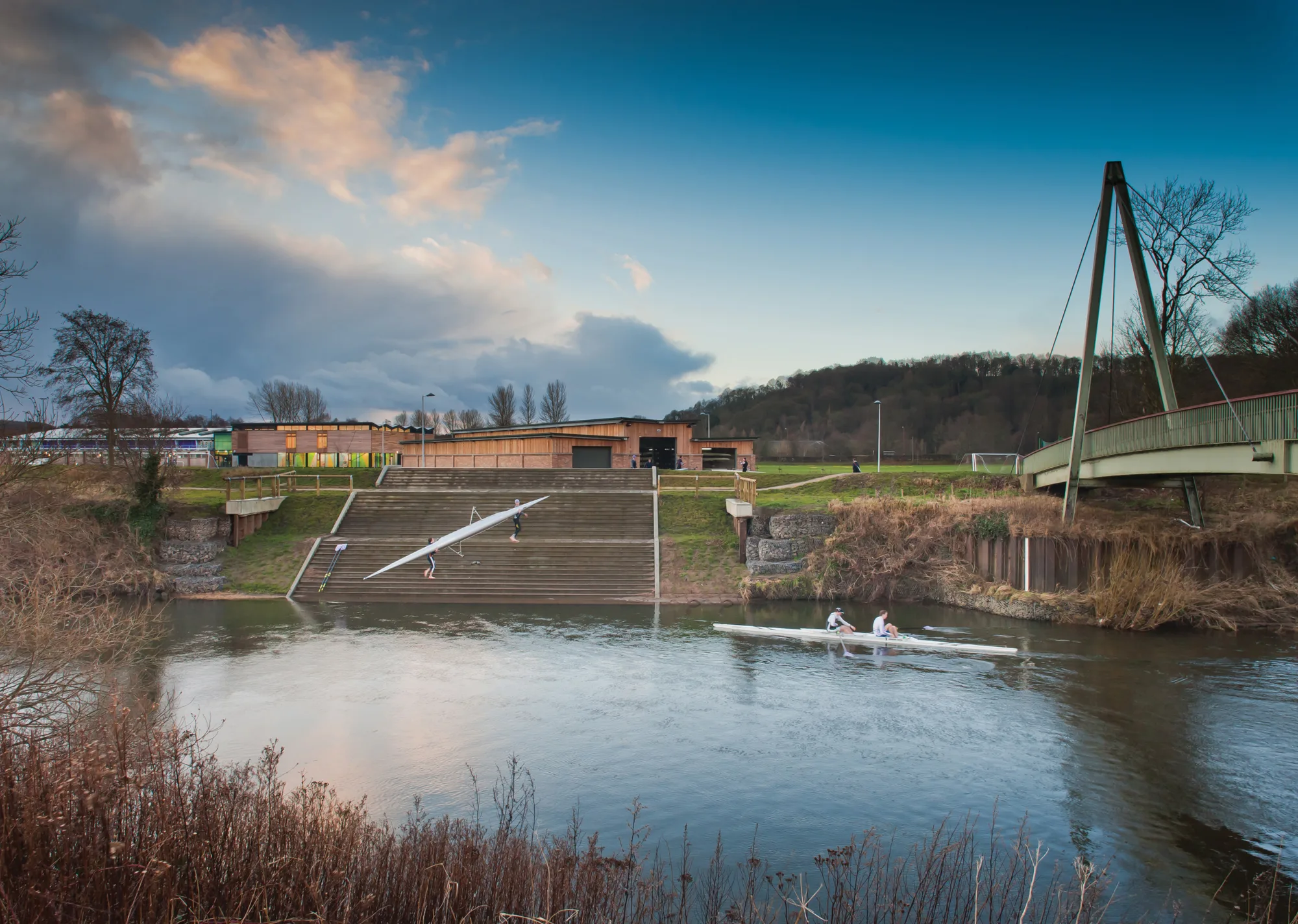 Wide river scene with a two‑person rowing boat in the foreground. Behind it are concrete steps leading to a wooden building, grassy banks, trees, and a suspension-style footbridge on the right.