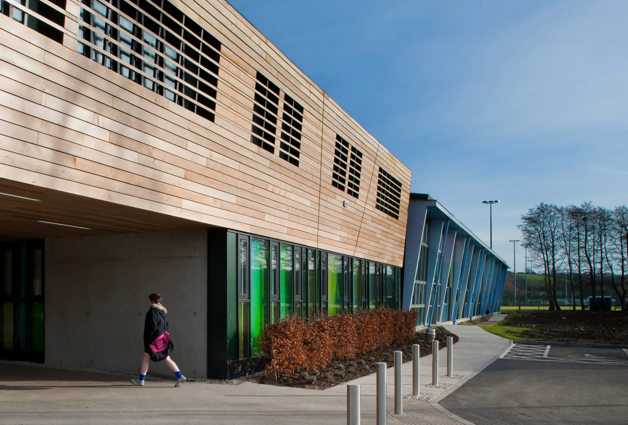 Side view of a modern building with wooden upper panels and green‑tinted lower windows. A person walks along the path beside shrubs and metal bollards, with a blue‑framed glass structure extending into the distance.