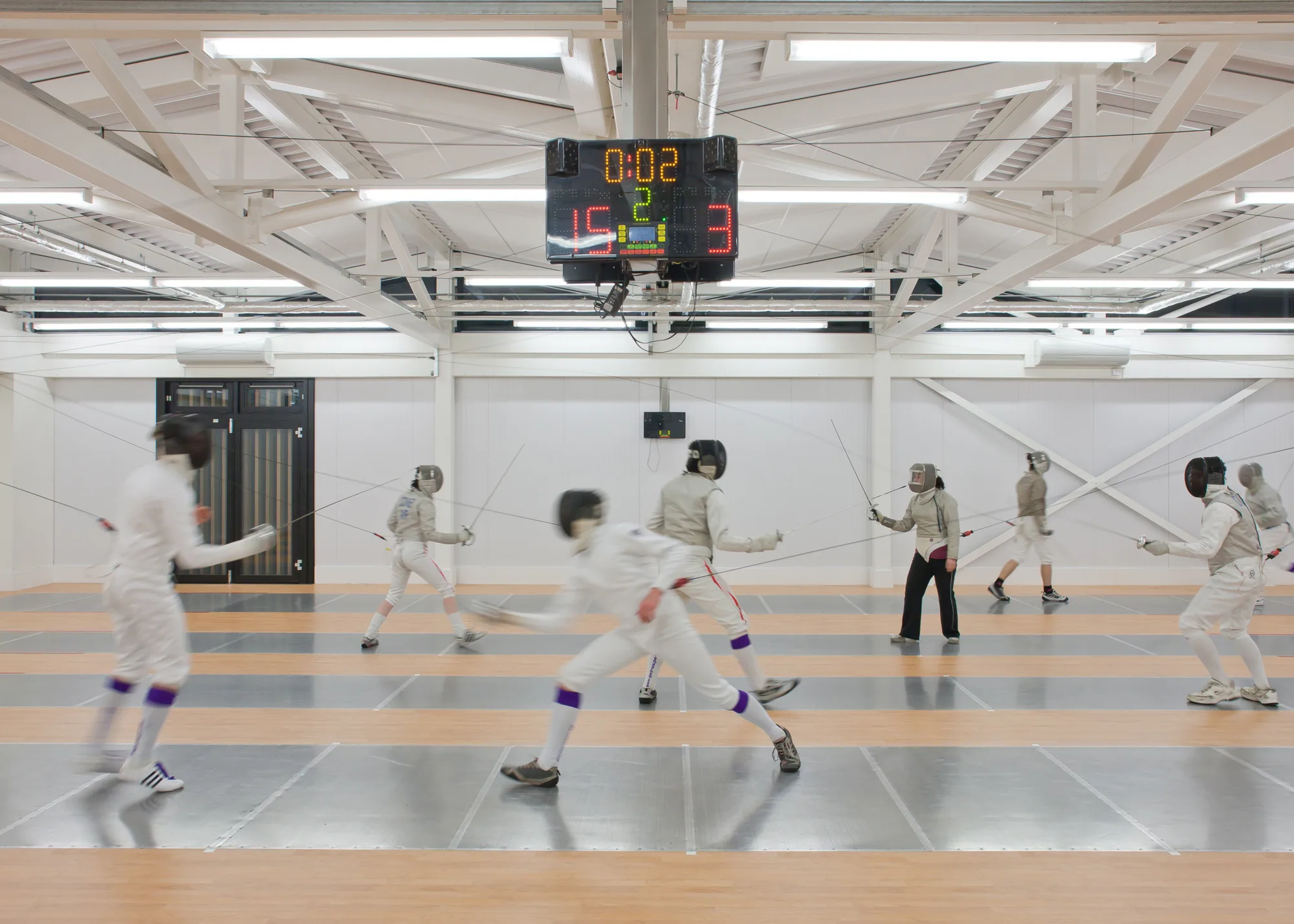 Indoor fencing hall with multiple people in full protective gear sparring on metallic pistes. A digital scoreboard above displays '00:02' and scores of 15 and 3. Bright overhead lighting illuminates the space.