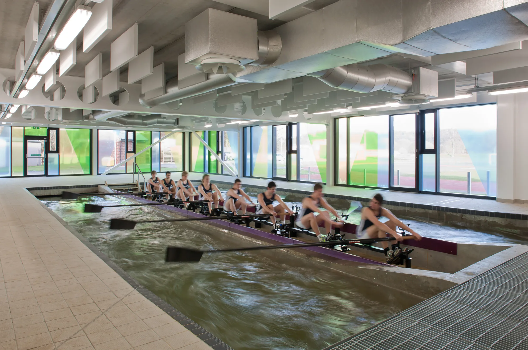 Indoor rowing tank with a long rowing platform holding multiple rowers using oars. Water flows beneath the platform, with overhead ducts and large windows along the wall in the background.