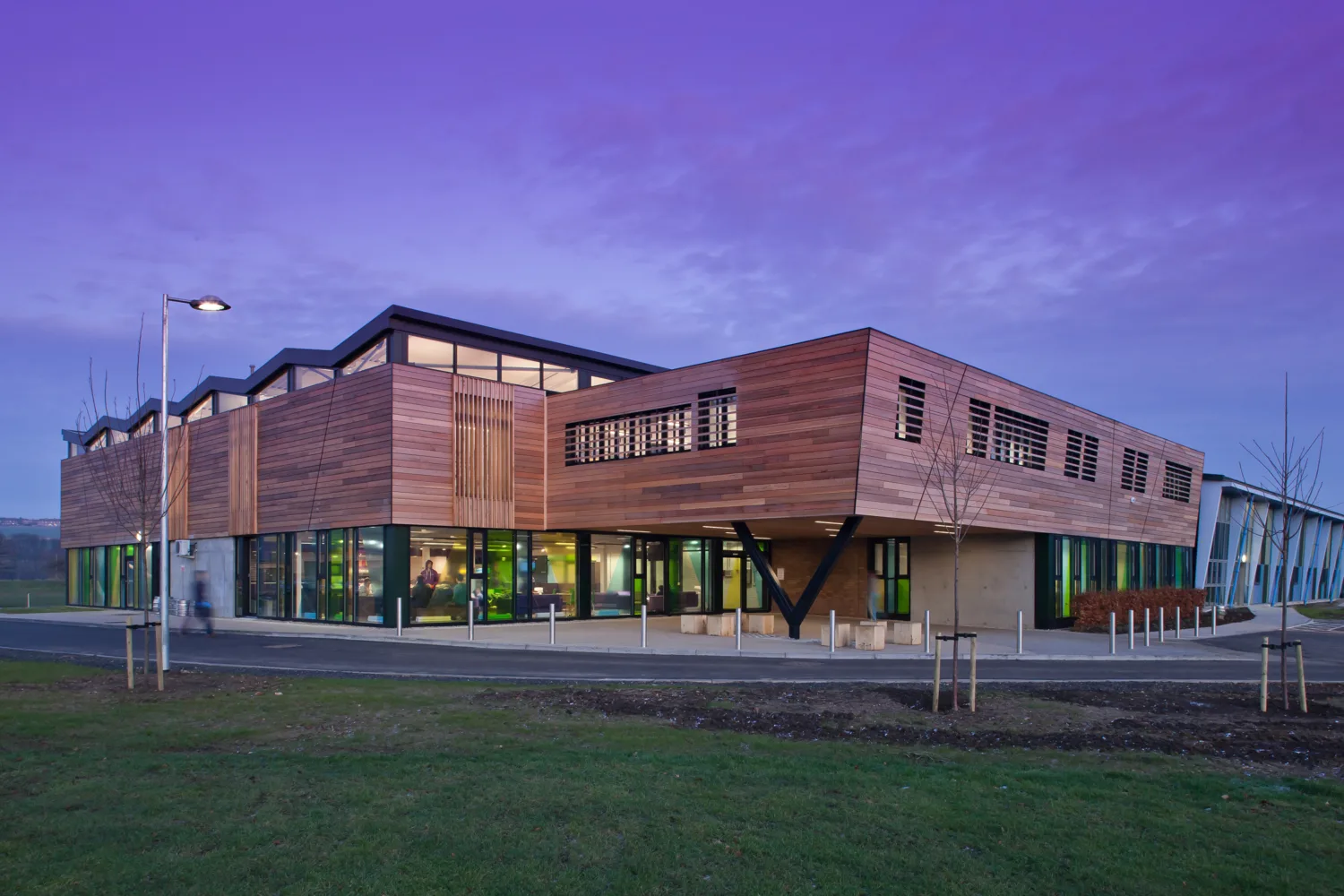 Modern two‑storey building with wooden exterior panels, large windows, and angled supports, shown at dusk with a purple sky. Exterior lighting, paved paths, and grassy areas surround the structure.