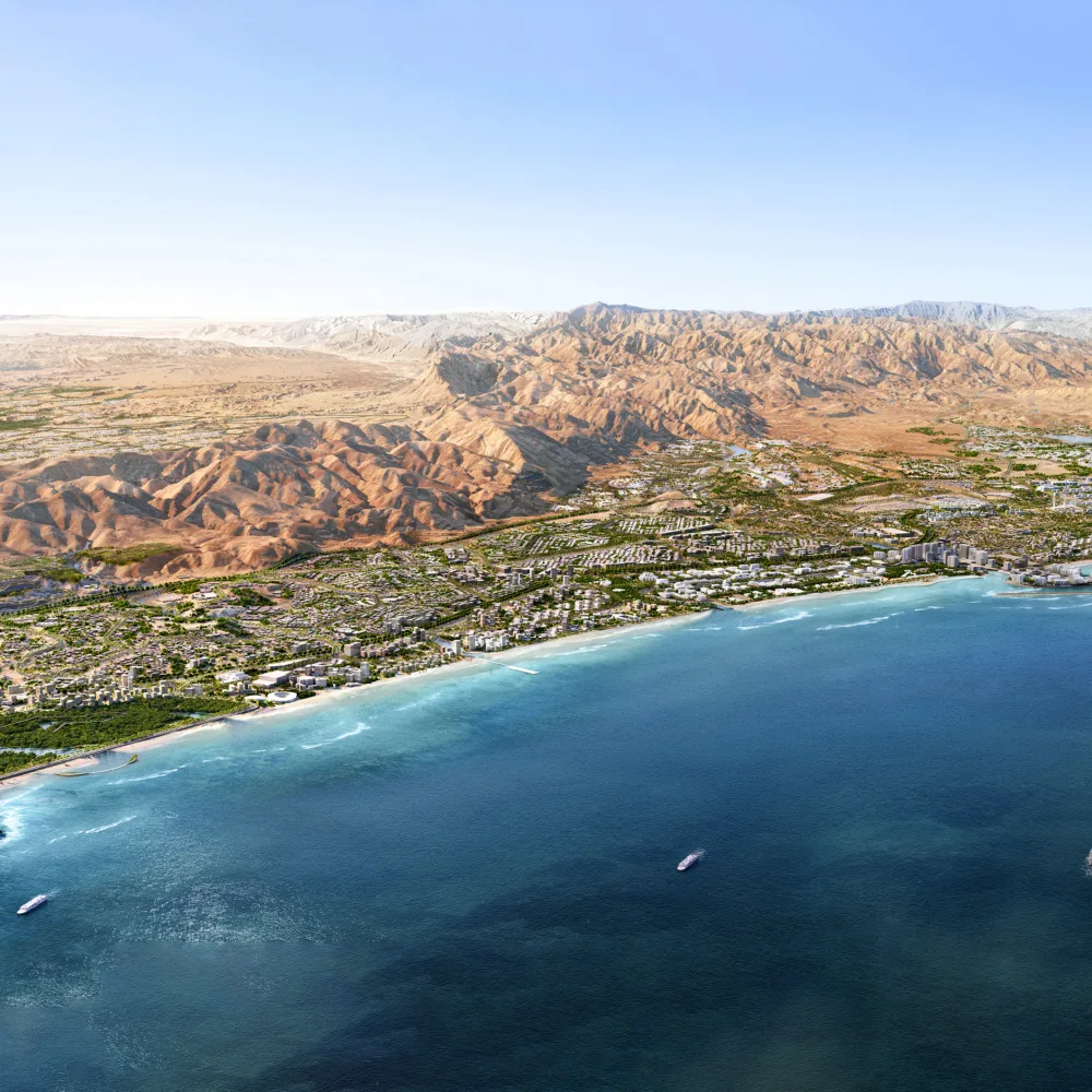 Aerial view of a coastal city with dense greenery, buildings, and roads along a curved shoreline. Clear blue water fills the foreground, and a wide expanse of rocky, beige mountains stretches across the background.