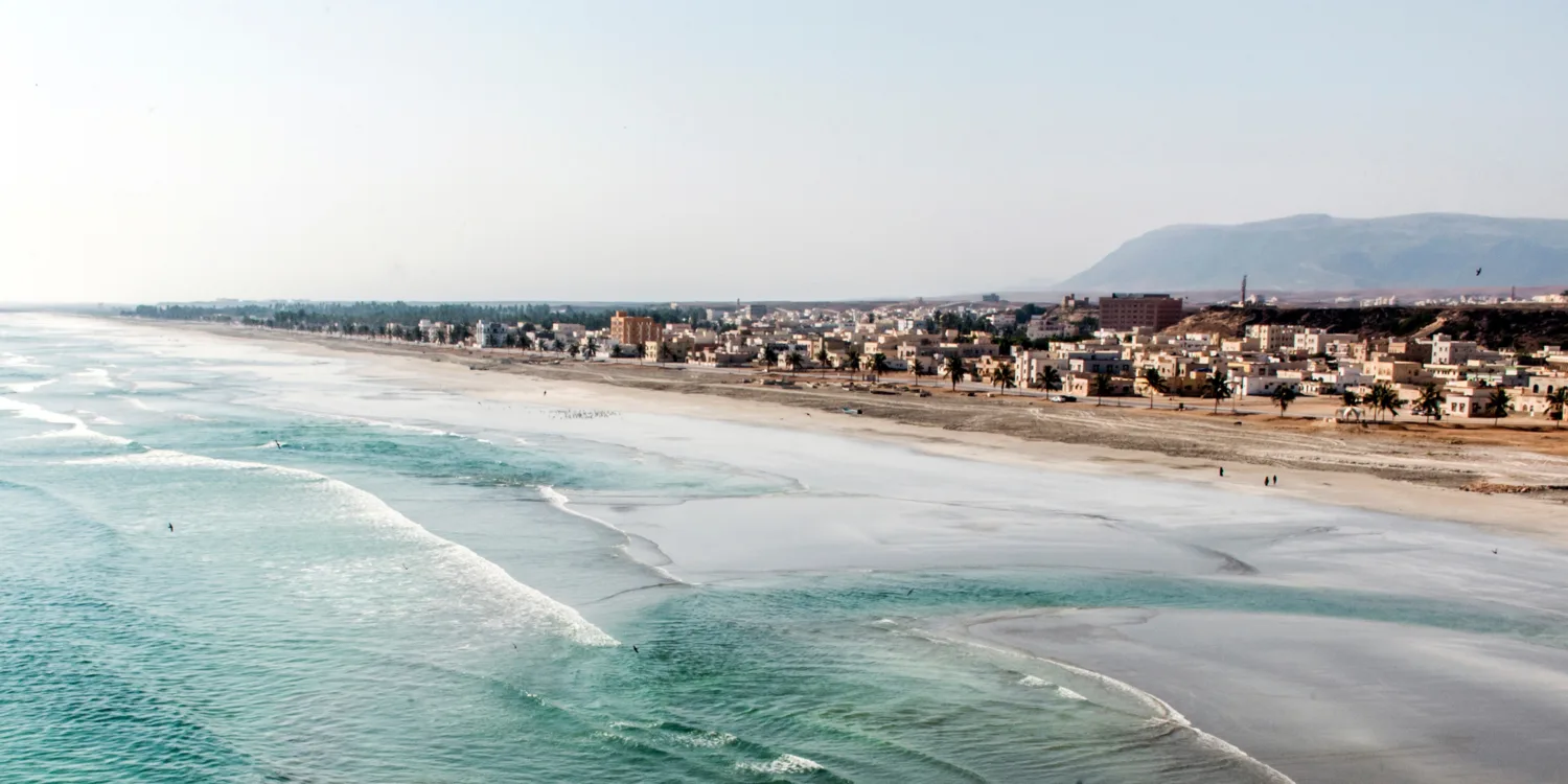 Wide coastal view of turquoise waves meeting a sandy beach beside a town with low-rise buildings and palm trees; hills in the background under a clear sky, with a few people walking along the shore.