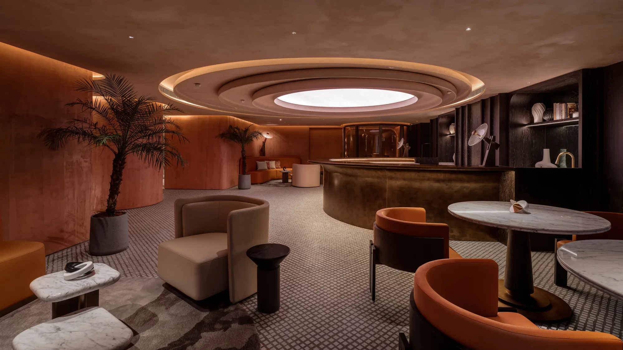 Modern lounge interior with curved copper-toned walls and ceiling, circular recessed light feature, marble tables, upholstered chairs, potted palm plants, and a rounded reception desk with shelves holding books and decorative objects.