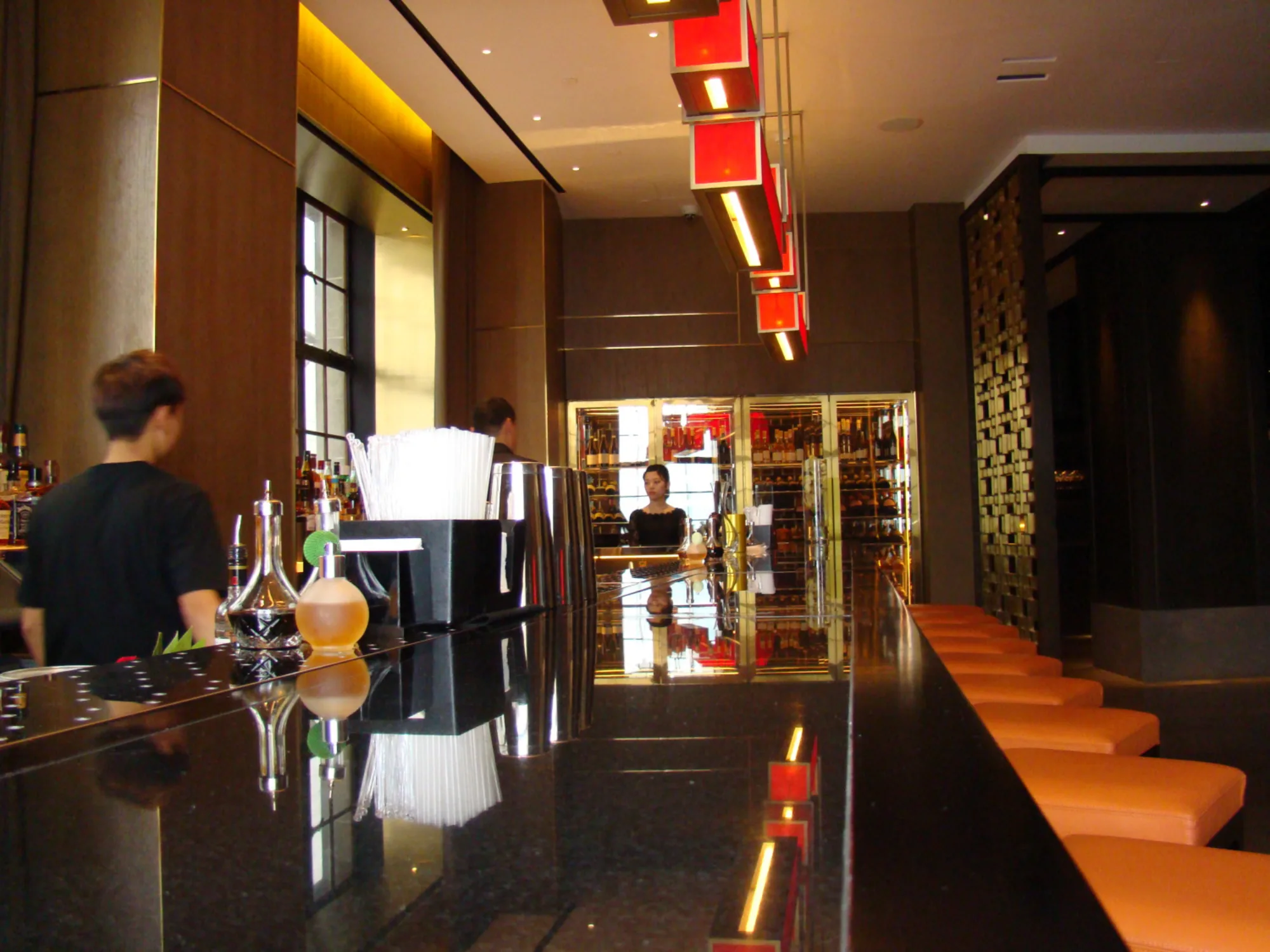 Restaurant’s bar featuring long countertop lined with bar stools, overhanging lights, and an array of drinks