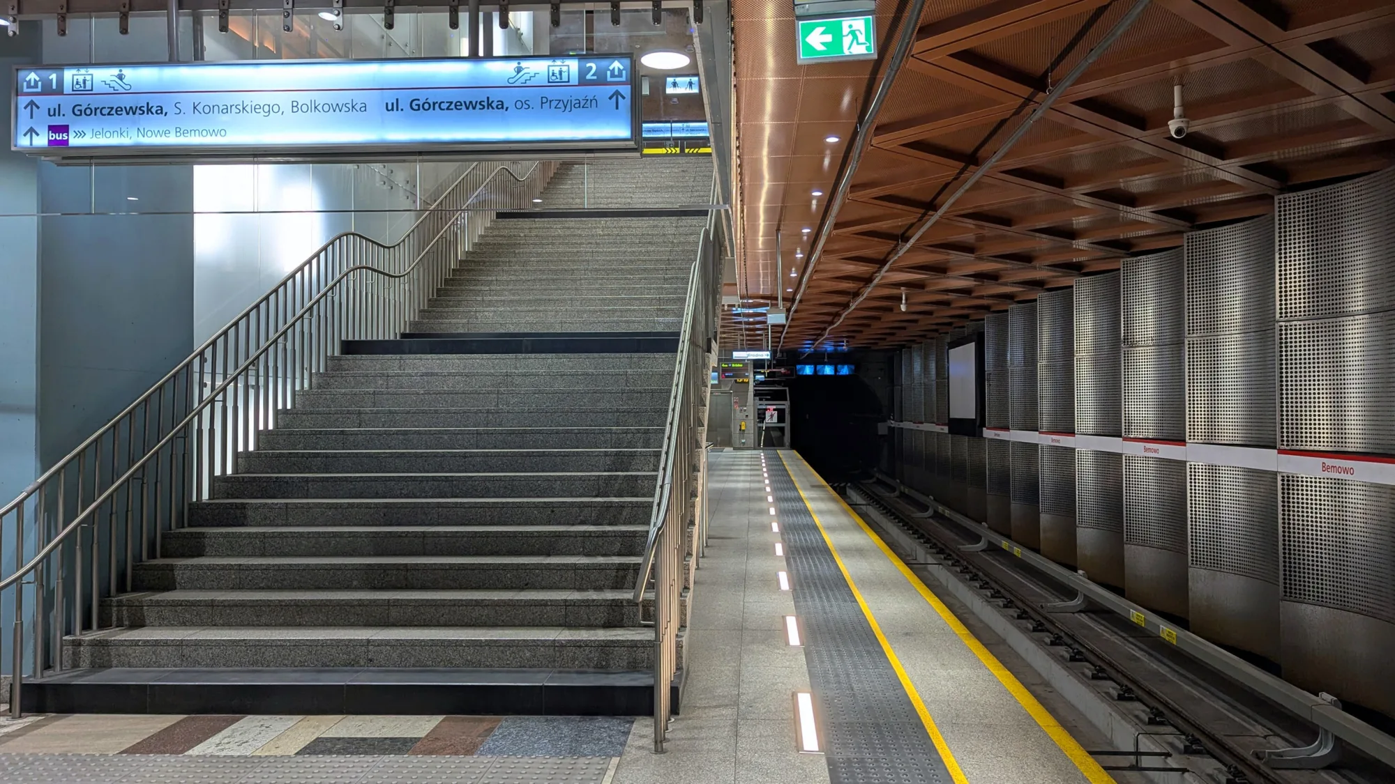 Concrete platform in the metro station separated from the tracks by 2 yellow lines leading into a tunnel. A staircase going up under illuminated sign.