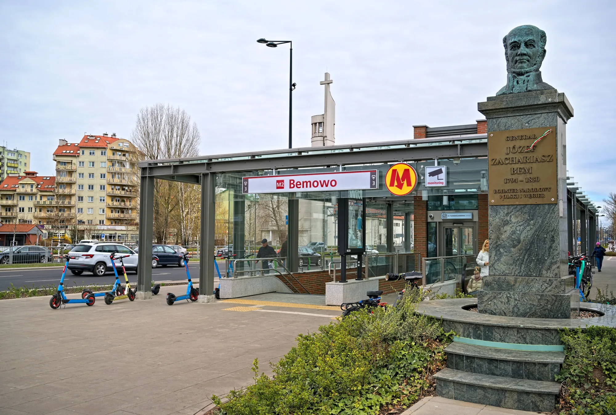 Exterior view of the metro station street level entrance with scooters parked at the top of the staircase and a statue to the right.