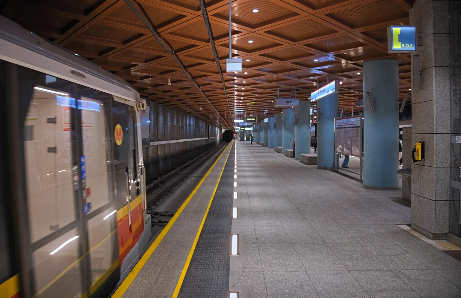 Platform view within the metro station as the metro enters. Yellow and white lines painted on the platform with a wooden ceiling.
