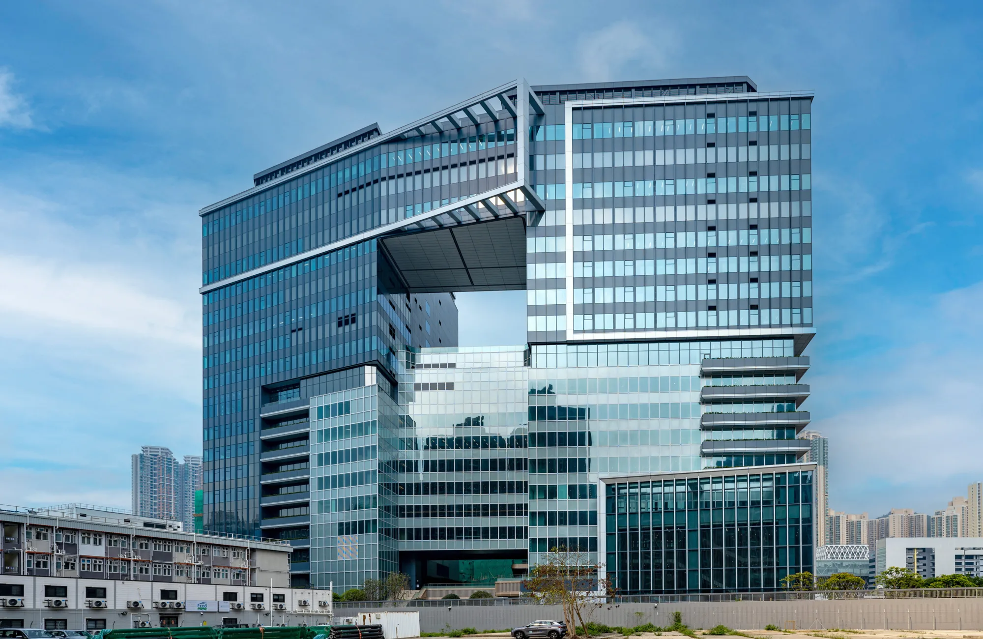 Multi-story glass office building with an open central section, reflective windows, blue sky background, construction site in foreground, and nearby low-rise buildings with greenery.