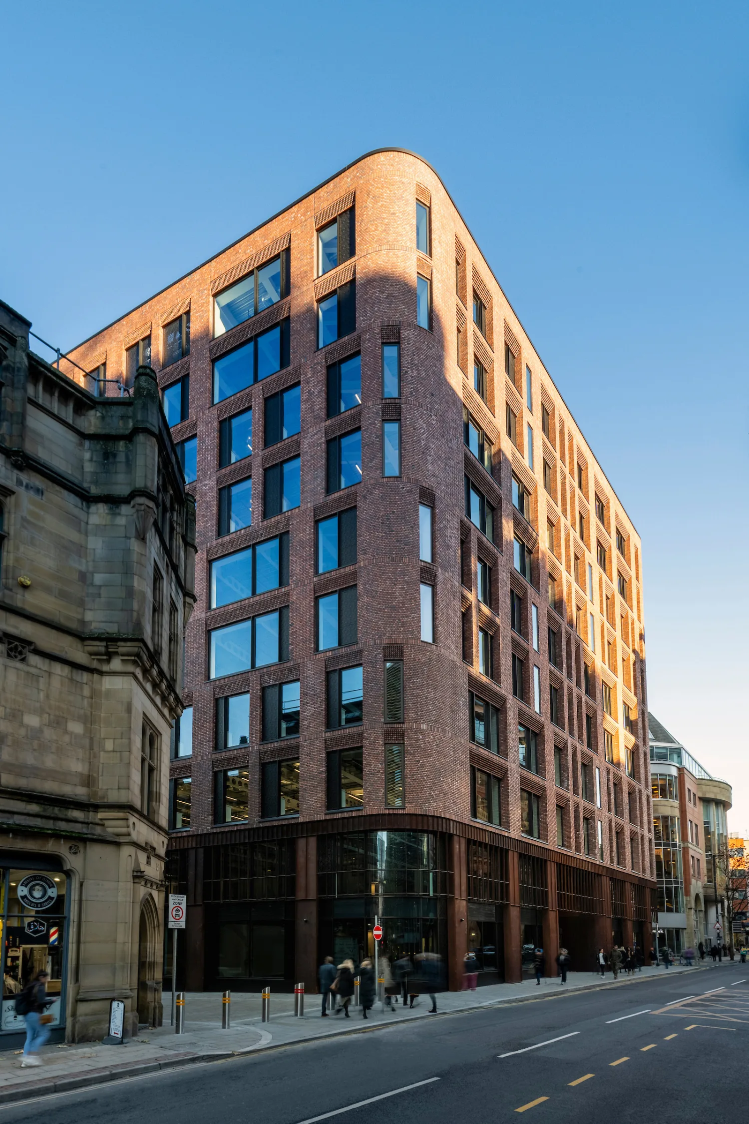 Multi-storey brick building with large windows and a curved corner next to an older stone building; clear blue sky, street signs, bollards, and pedestrians on the pavement.