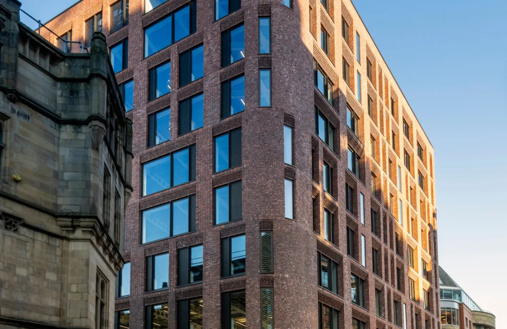 Multi-storey brick building with large windows and a curved corner next to an older stone building; clear blue sky, street signs, bollards, and pedestrians on the pavement.
