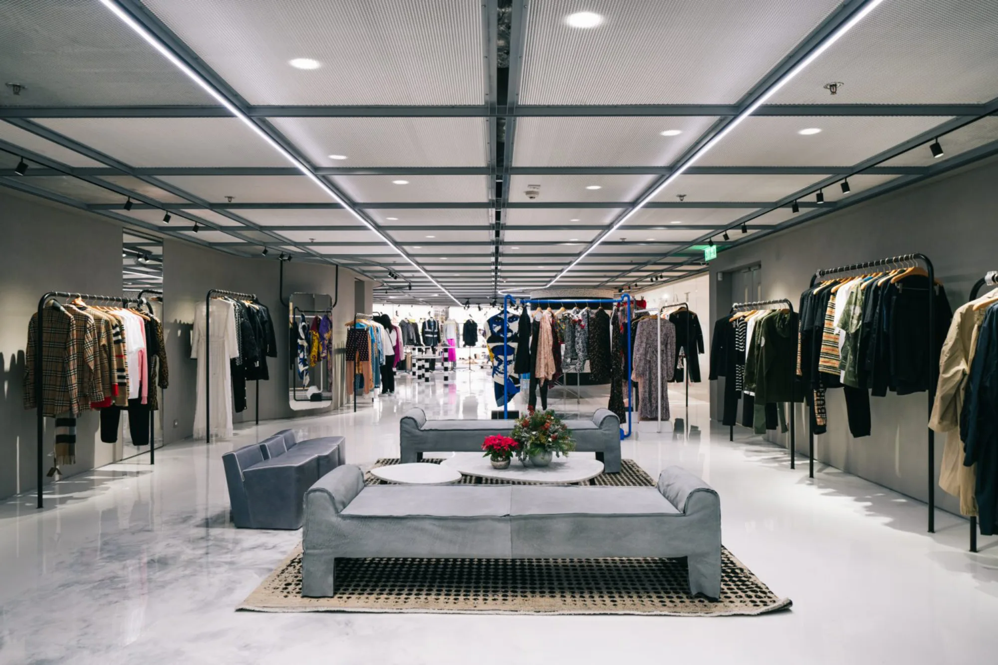Overview shot of the whole boutique with three sofas in the centre and clothing located on both sides. Tailor-made downlight fixture can be seen on the metal mesh ceiling.