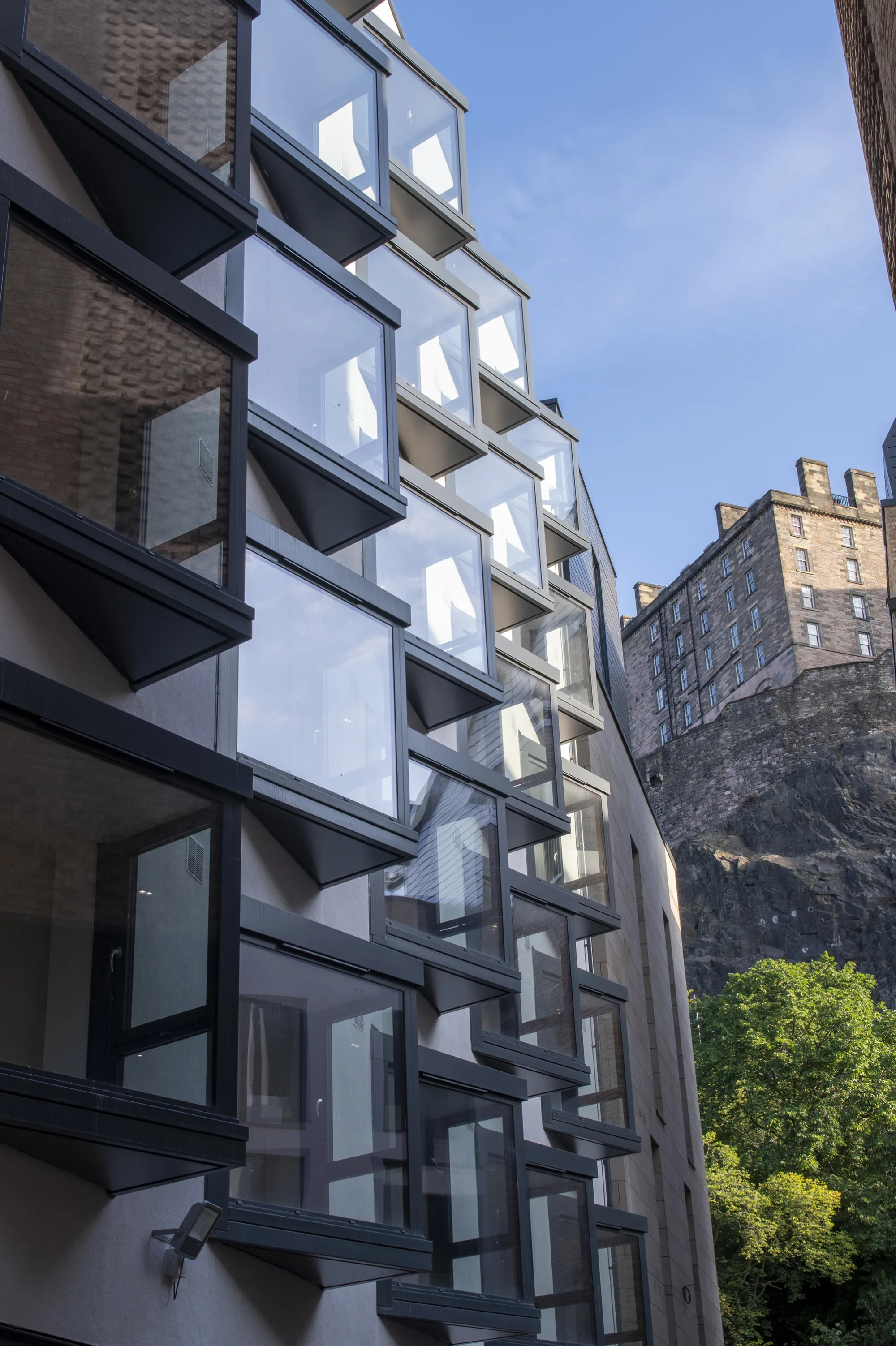 Edge of building with jutting windows in forefront with Edinburgh Castle in background, set against blue skies.
