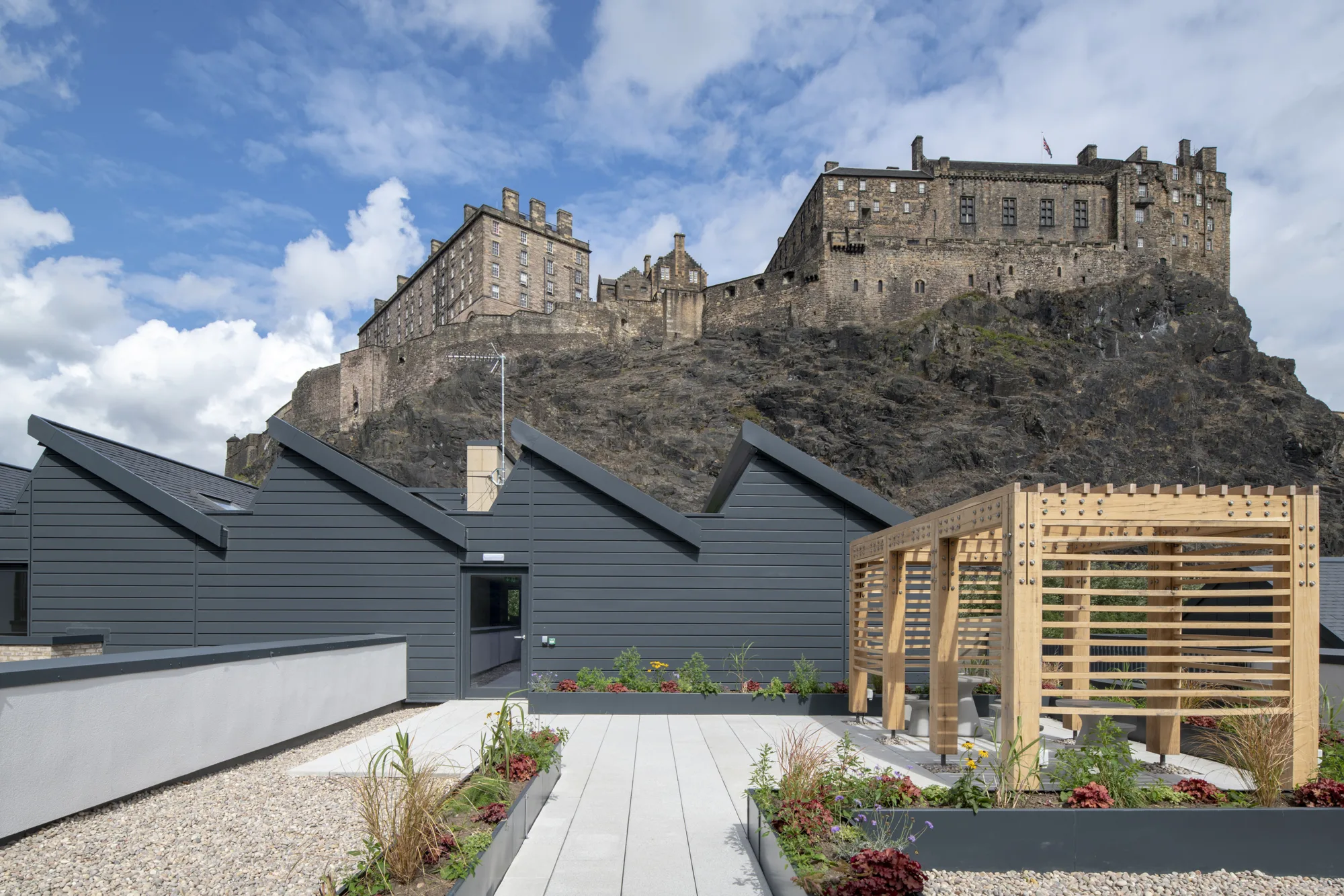 Edinburgh Castle in background, with gravelled patio including sheds, flowerbeds and wooden structure in foreground