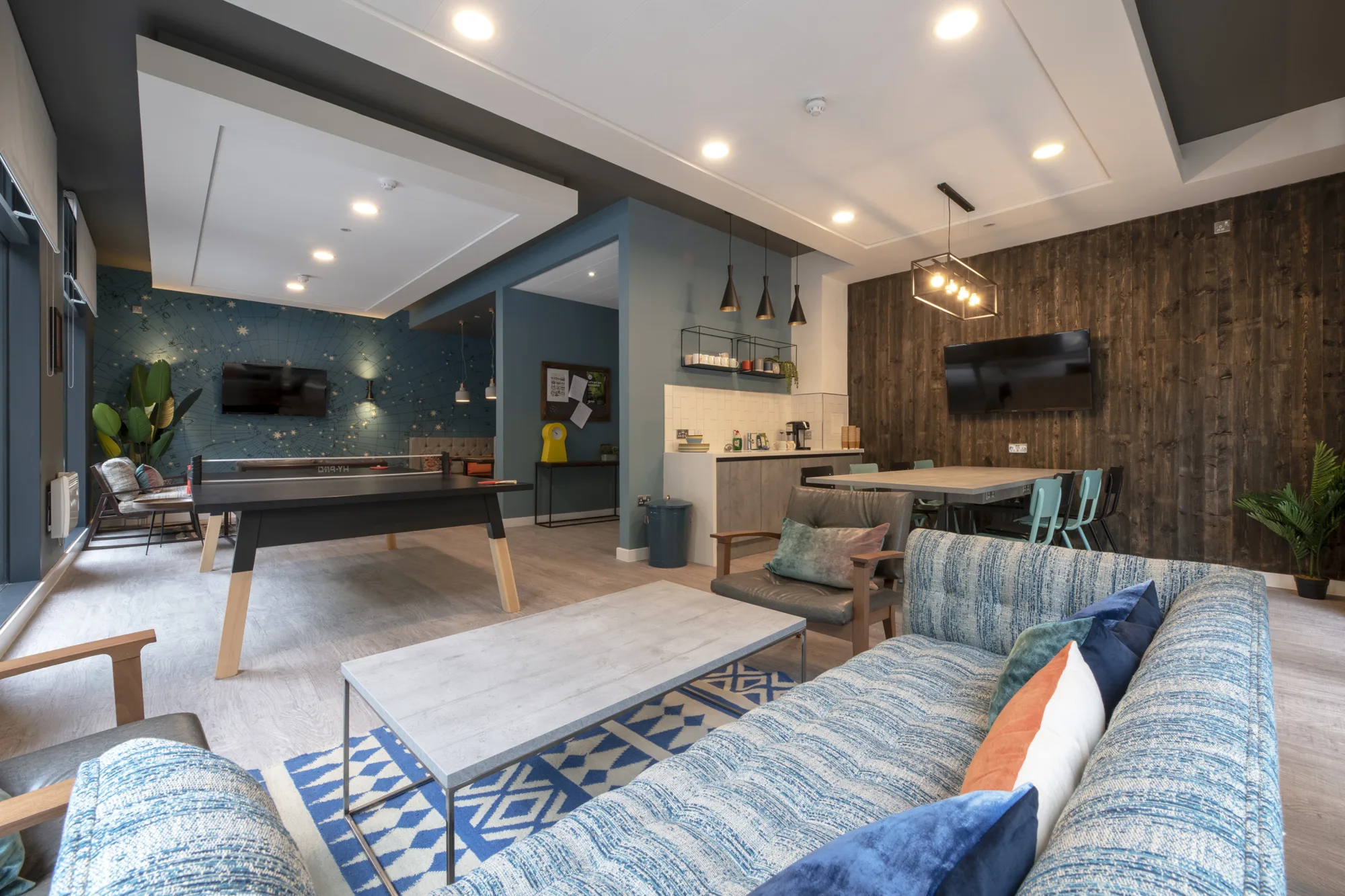 Common room with blue walls including sofa, pool table, kitchen area, table and chairs, TVs and innovative lighting.