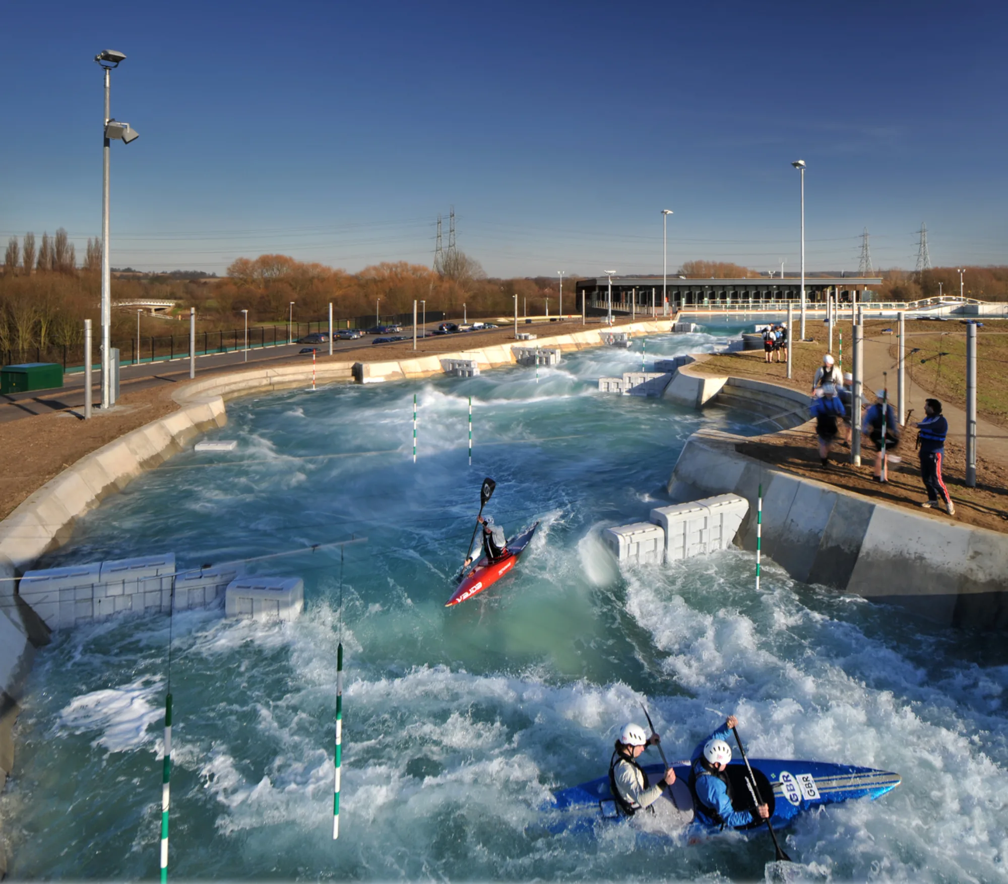 A man‑made white‑water course with fast flowing water, gates, and concrete barriers. Several kayakers navigate the rapids while a few people stand on the bank near fencing and equipment.