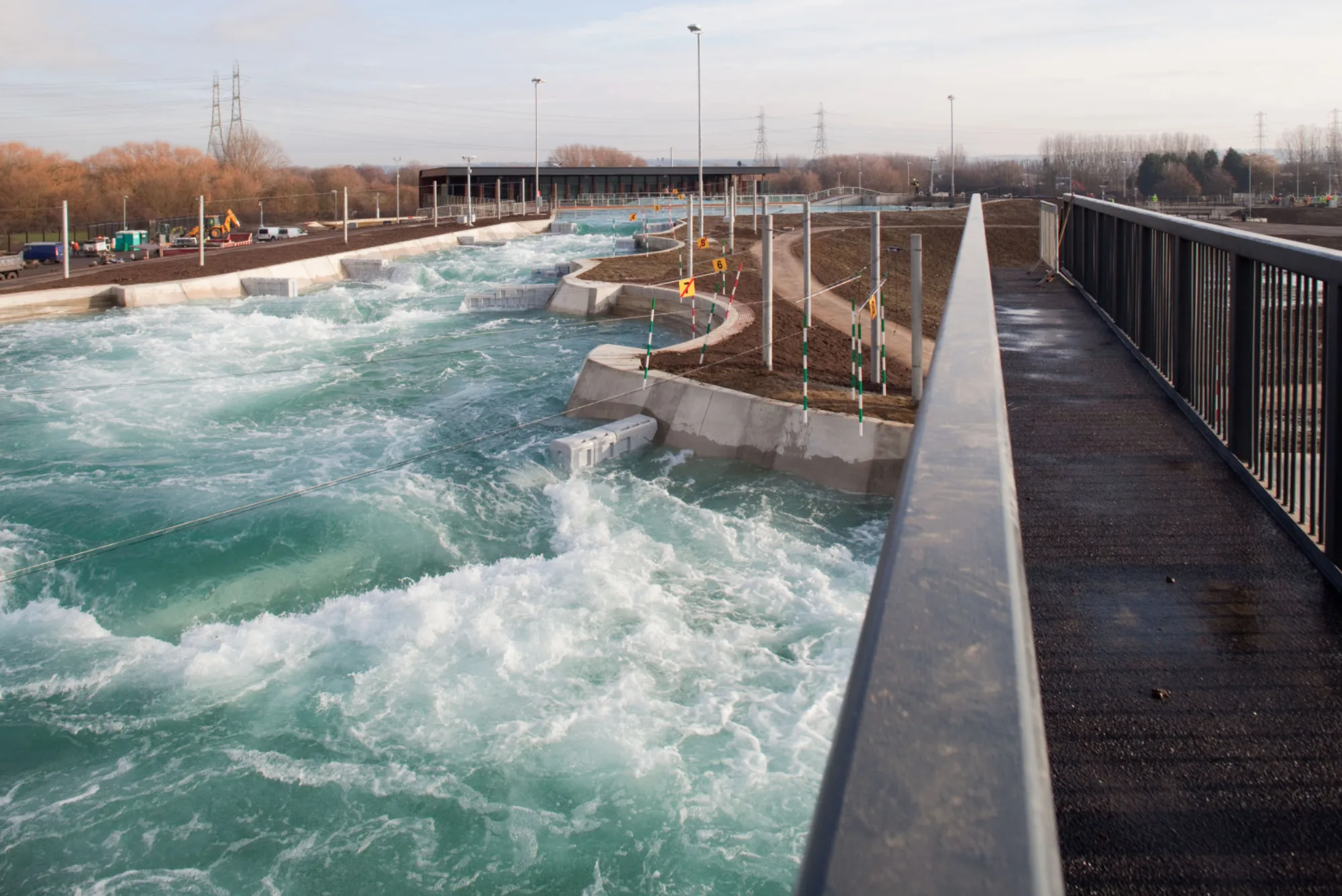 Outdoor white‑water course with fast flowing water, concrete channels, and slalom poles. A walkway with metal railings runs alongside, with buildings, vehicles, and power lines in the background.