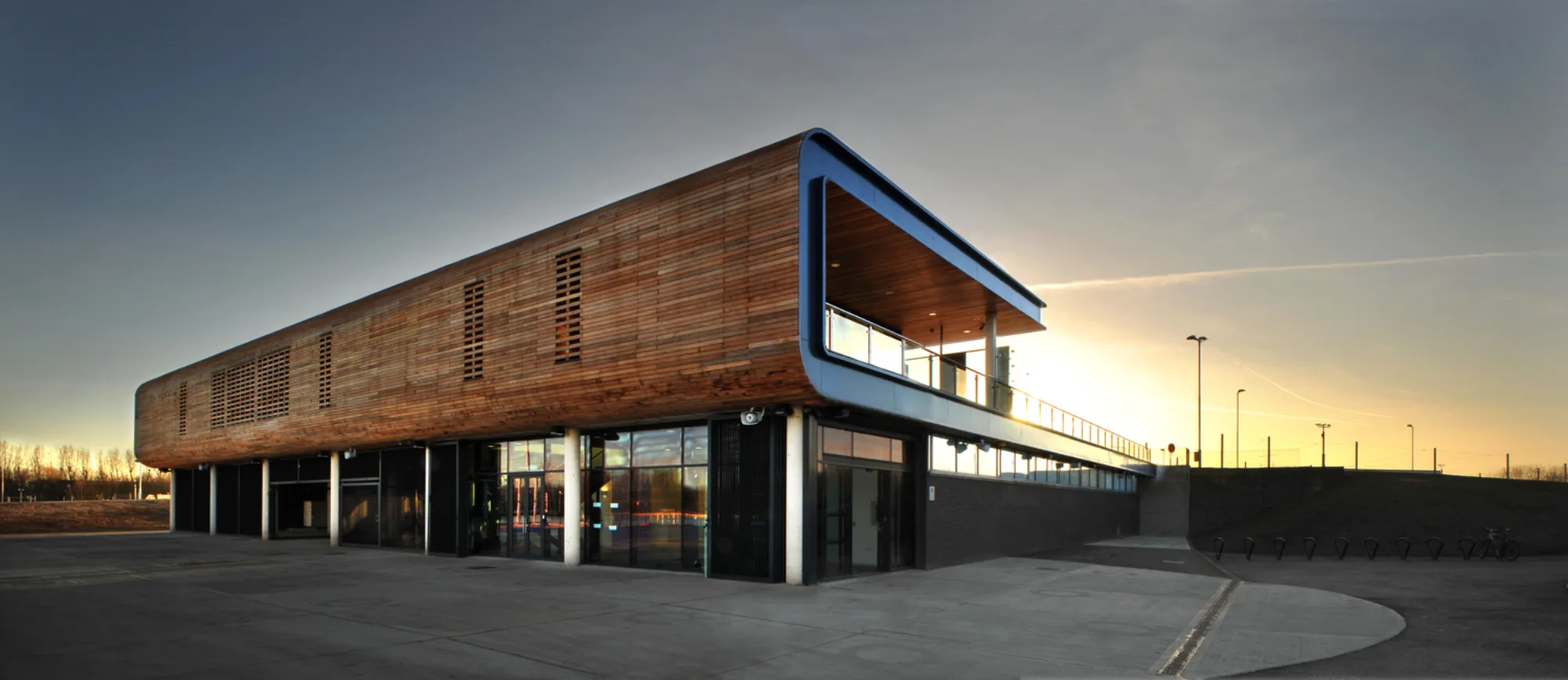 Modern two‑storey building with a wooden upper facade, large glass windows, and metal supports, shown at sunset on an open paved area with bike racks and tall light poles nearby.