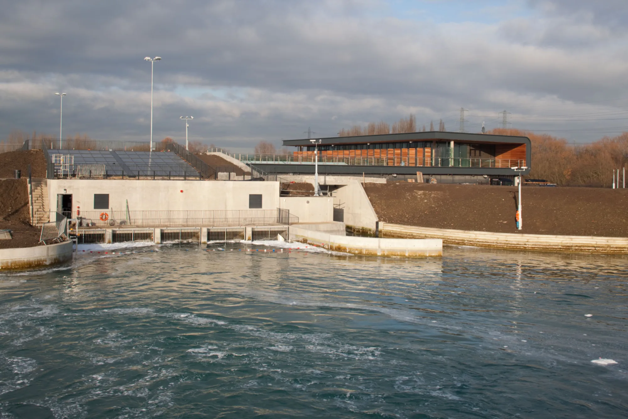 Large outdoor water facility with a concrete structure, railings, and churning water in the foreground. A modern two‑storey building with wood and glass sits on a raised bank under a cloudy sky.