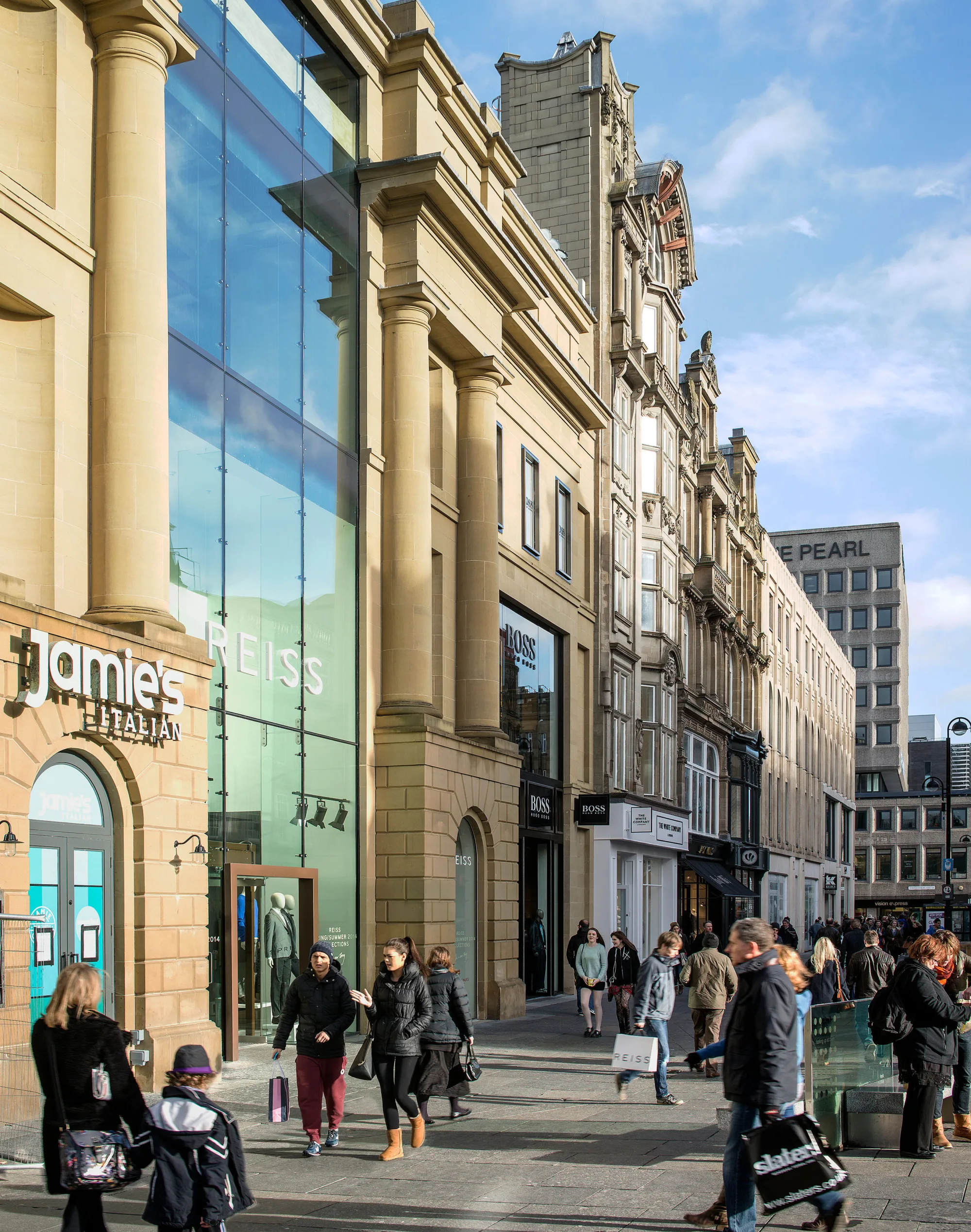 Street-level view of sandstone buildings with large glass storefronts. Visible signs include ‘jamies ITALIAN,’ ‘REISS,’ and ‘BOSS.’ Several people walk along the pavement carrying shopping bags under a clear sky.