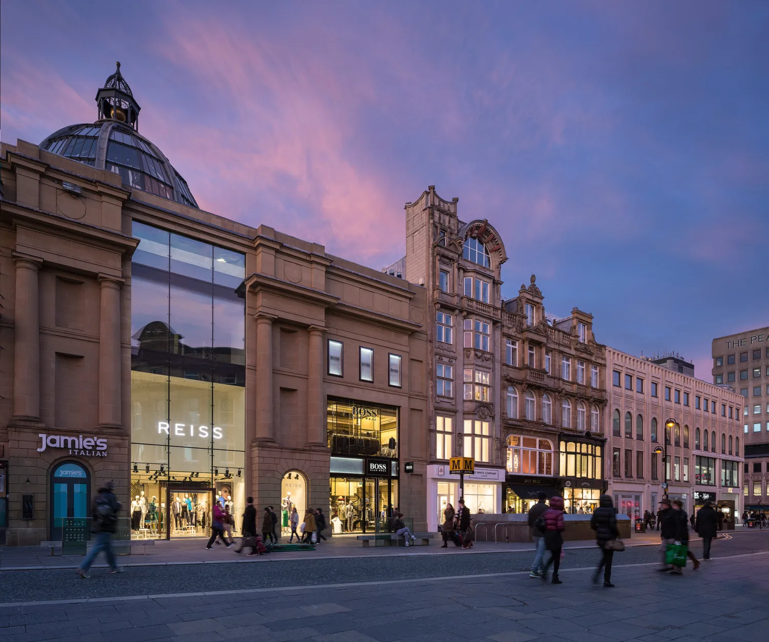 Street view of Monument Mall at dusk with a pink and purple sky. Large sandstone buildings with glass storefronts display signs for ‘REISS,’ ‘jamies,’ and ‘McDonald’s.’ Several people walk along the cobblestone pavement.