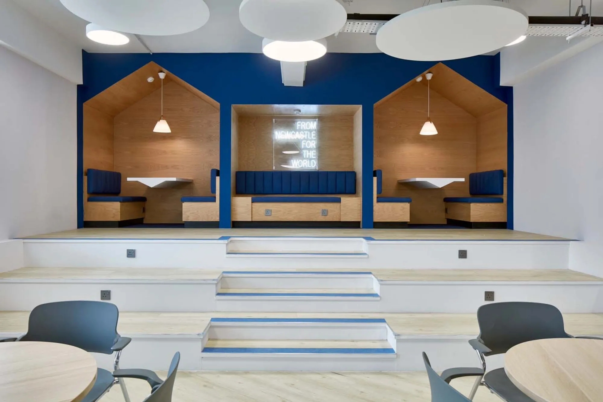 Brightly lit workspace with round tables and platform / wide steps at the back leading up to three work booths
