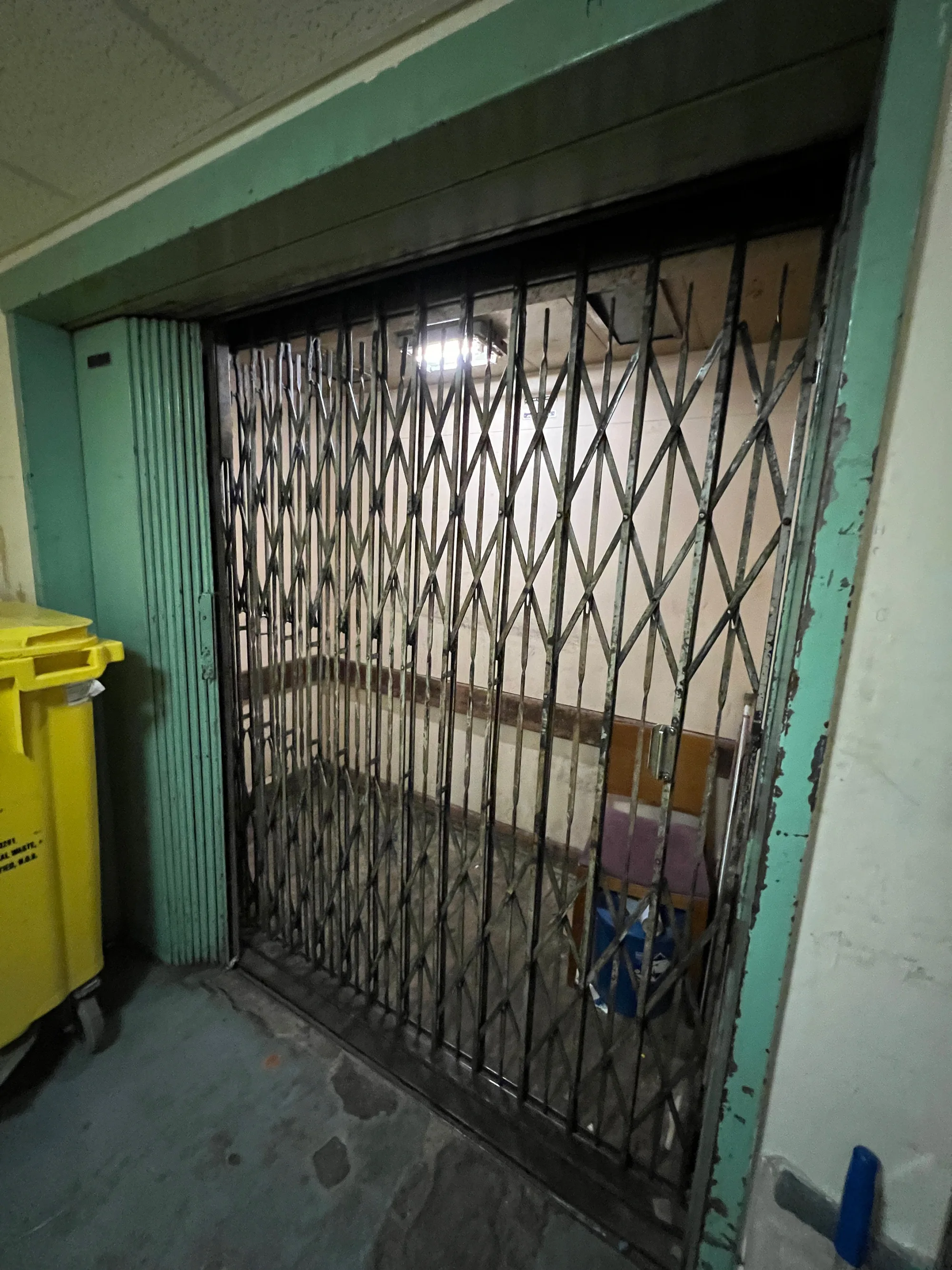 An open elevator with a metal accordion gate, green frame, and dim interior. A yellow waste bin sits to the left, and various objects are visible inside the lift area.