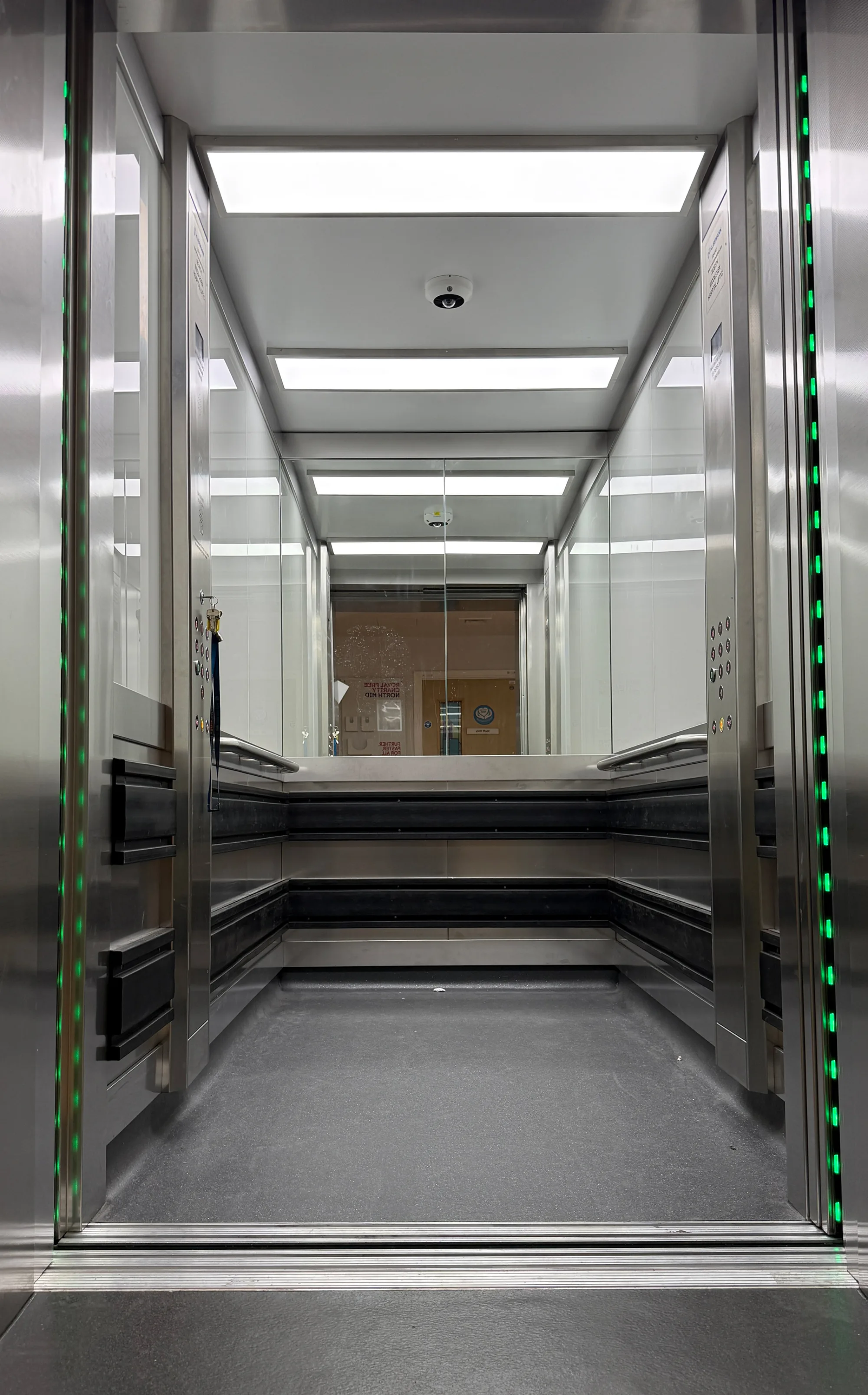 An open modern elevator with mirrored walls, bright ceiling lights, handrails on three sides, control panels on both door frames, and green indicator lights along the door edges.