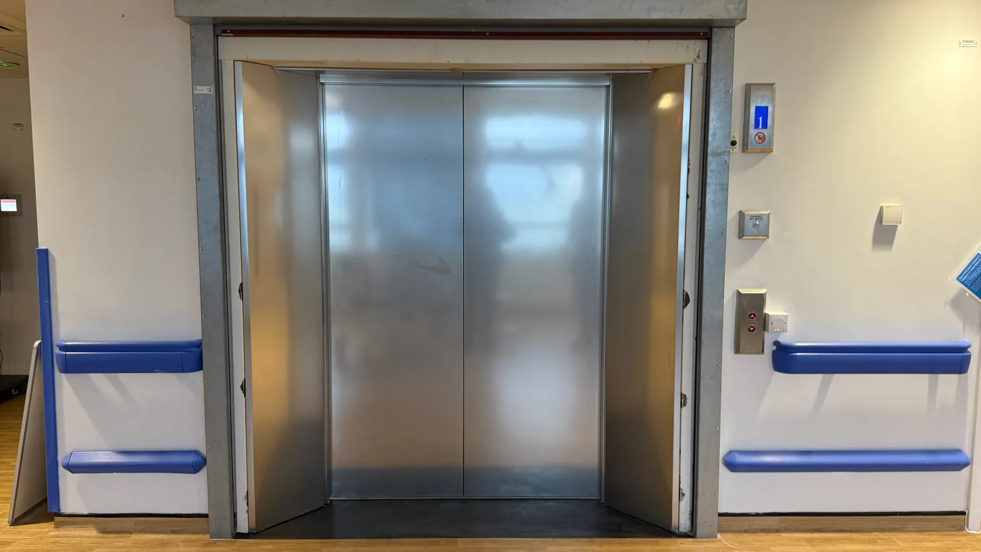 Closed stainless‑steel elevator doors in a hospital corridor, with blue wall‑mounted handrails on both sides and a control panel to the right displaying an illuminated blue floor indicator.