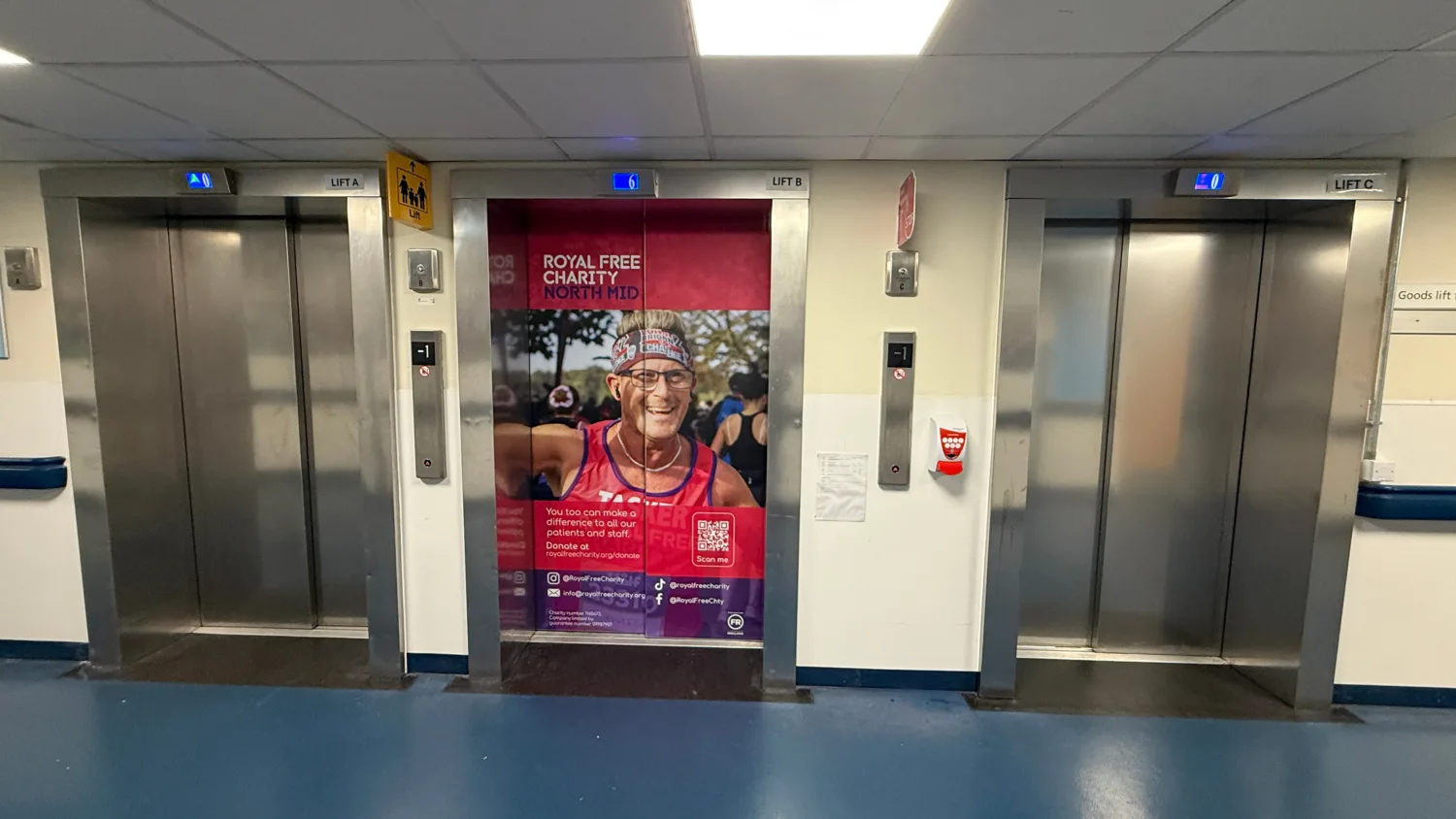 A hospital corridor with three elevators; the middle elevator door displays a large poster showing a runner in a pink vest with text reading ‘Royal Free Charity North Mid’ and a QR code below.