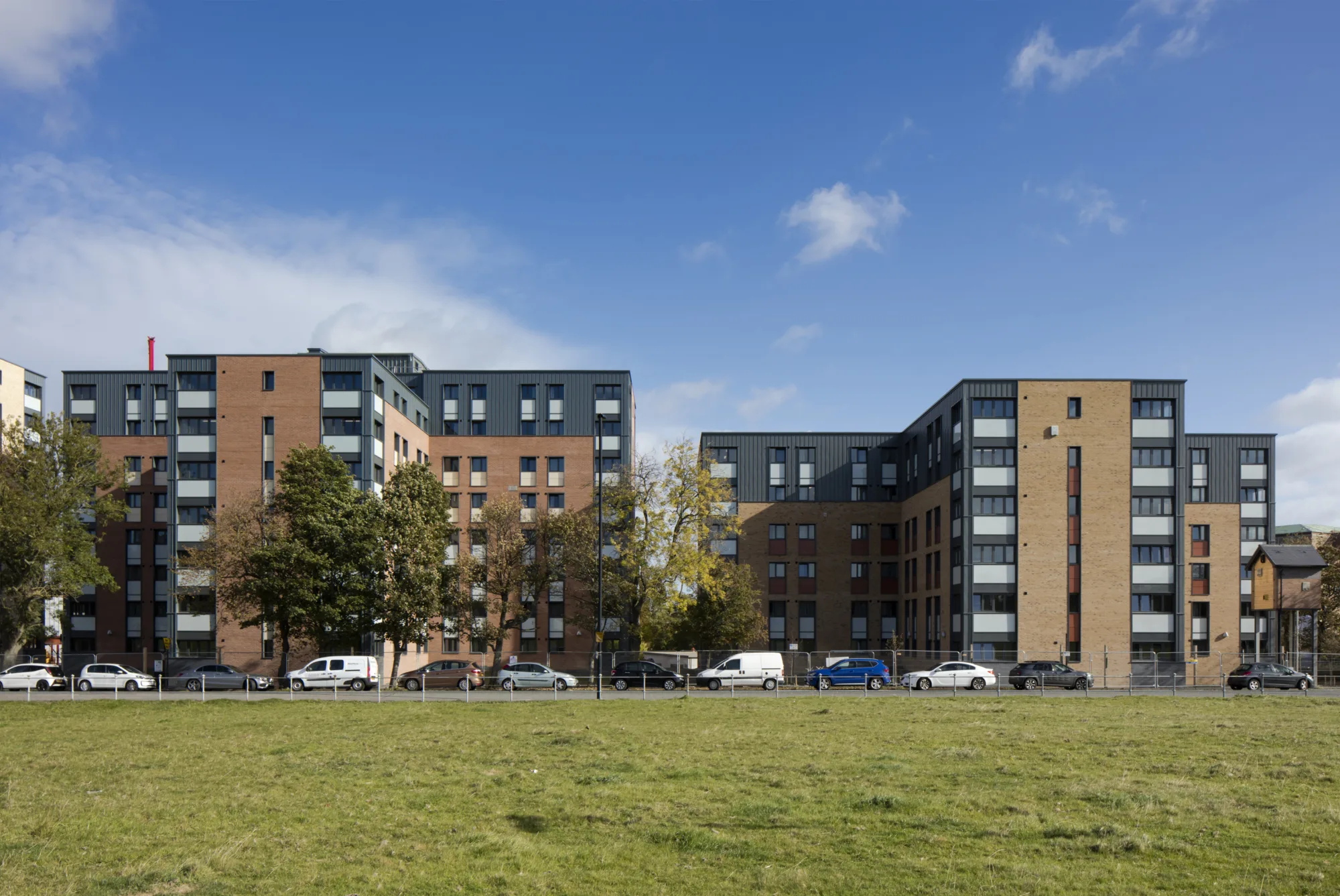 Straight-on view of the front of two student village buildings with dark grey and light brown exteriors. In front is a road with parked vehicles and a grassy field.