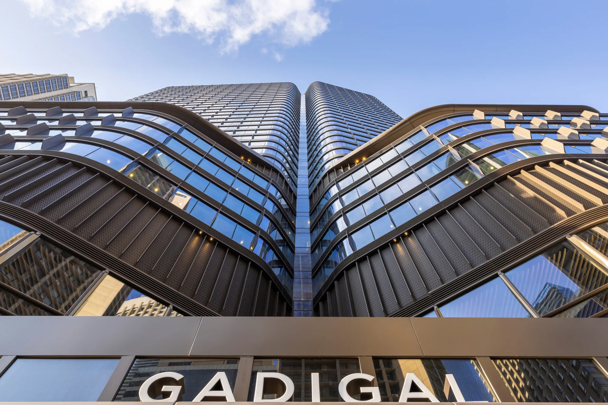 Low-angle view of a modern high-rise building with curved, layered glass and metal façade reflecting the sky and nearby structures. The word “GADIGAL” is visible on a sign at the bottom of the image.