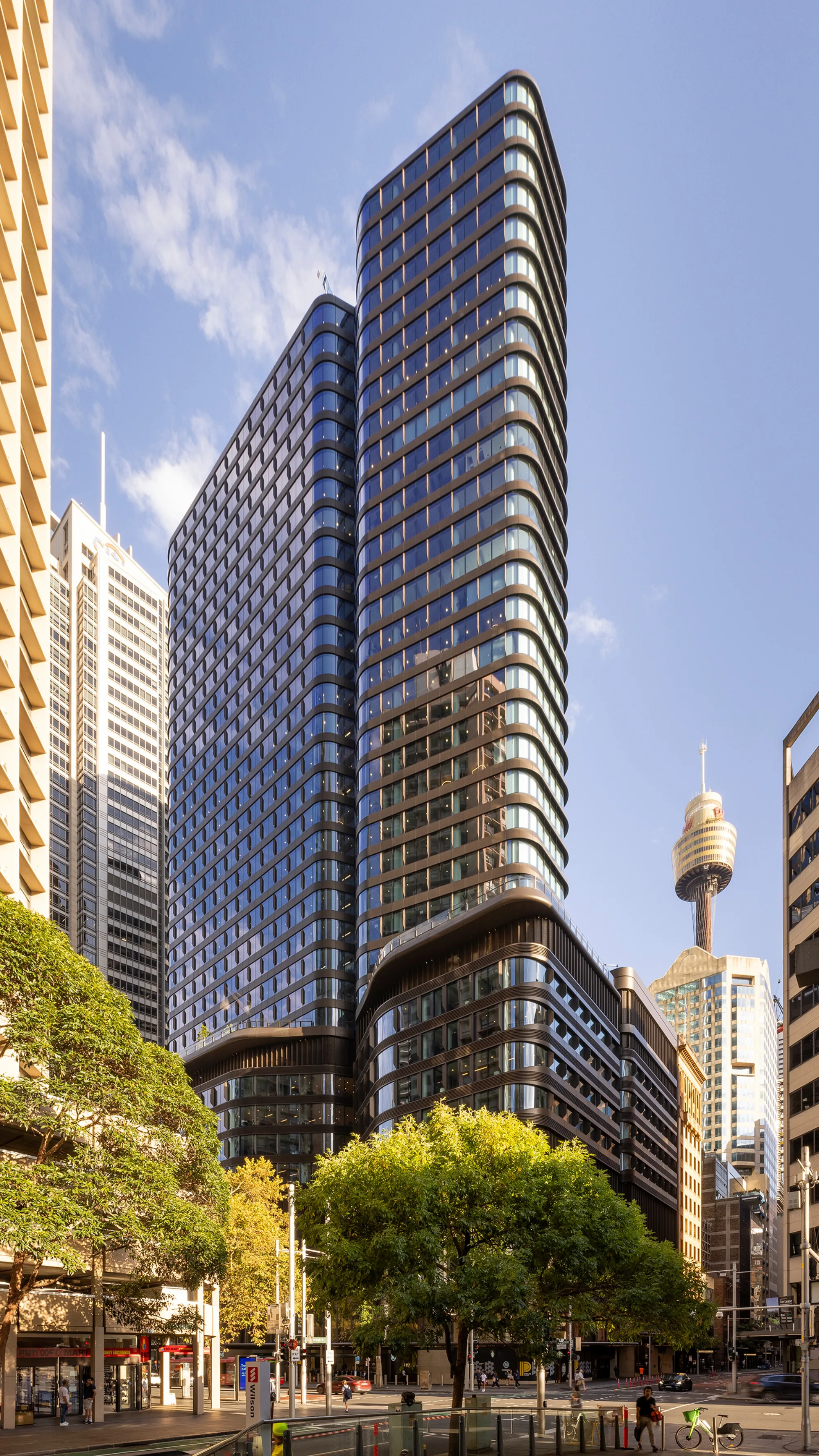 Tall modern glass building with curved edges rising above surrounding structures in a cityscape. Trees and street-level traffic are visible in the foreground, with a clear blue sky in the background.