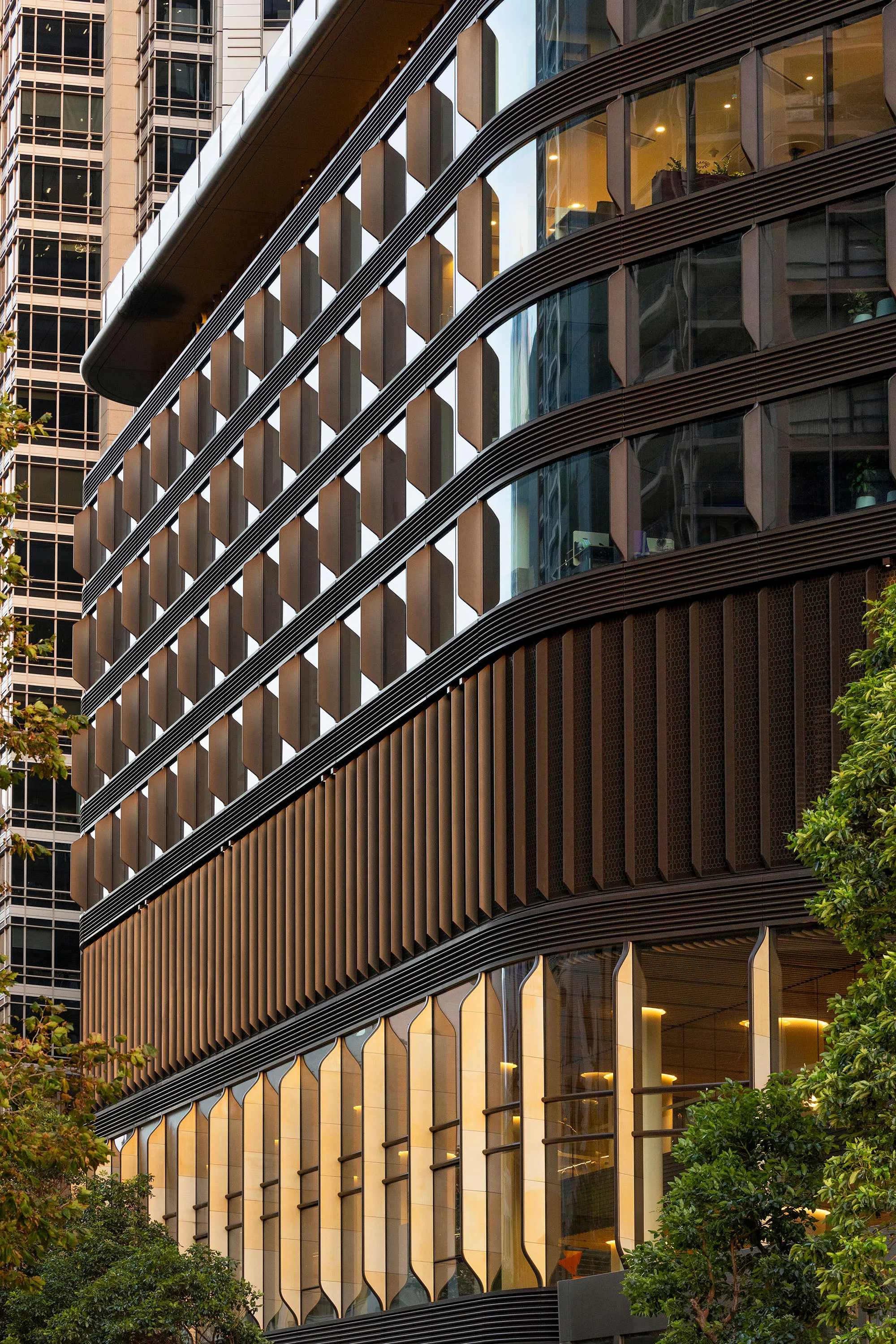 Close-up view of a modern building façade featuring horizontal metal fins, large glass windows, and a lower section with tall vertical panels. Trees partially frame the foreground.