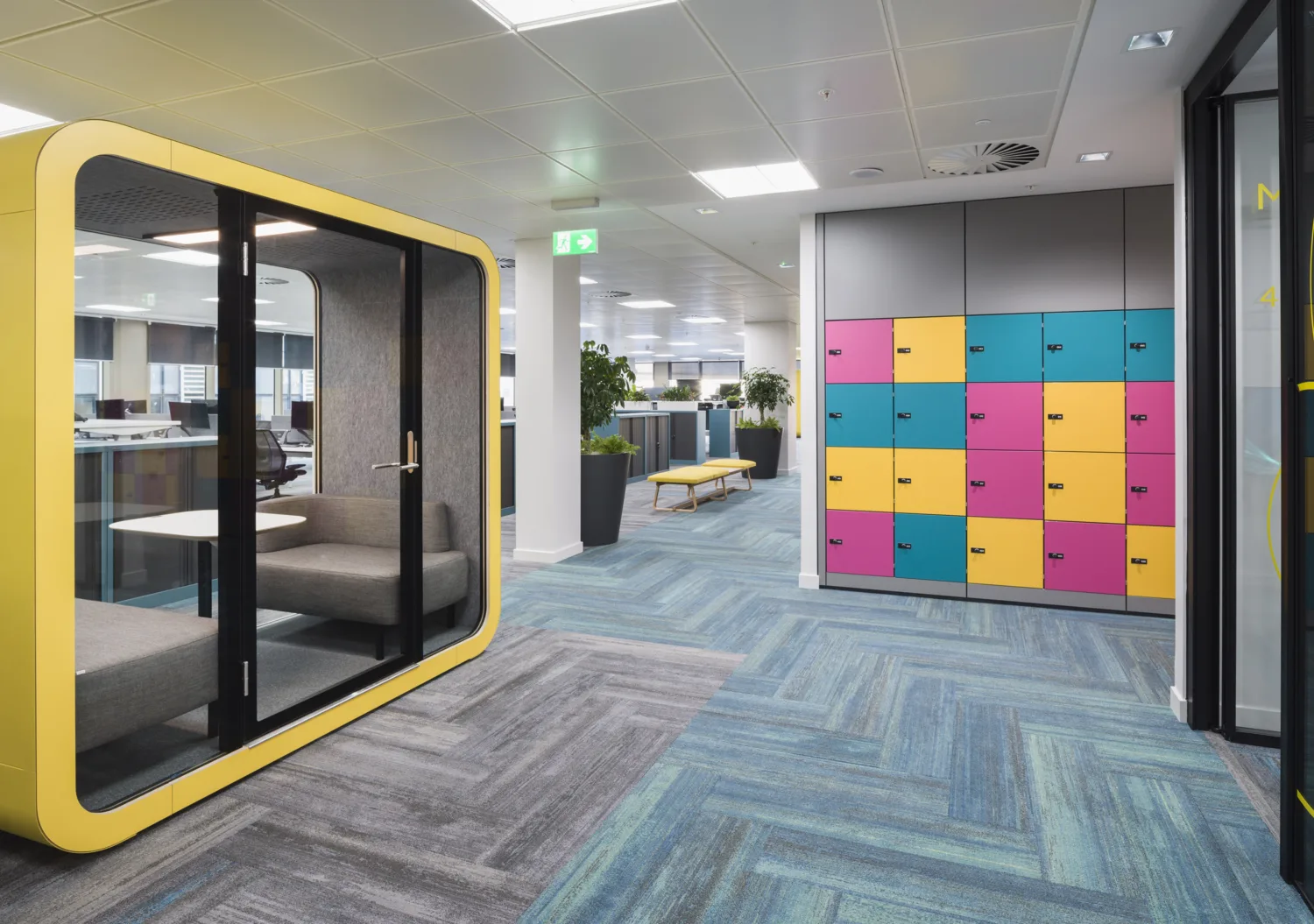 Office interior with blue and grey carpet, storage lockers and a yellow hub glassed meeting area