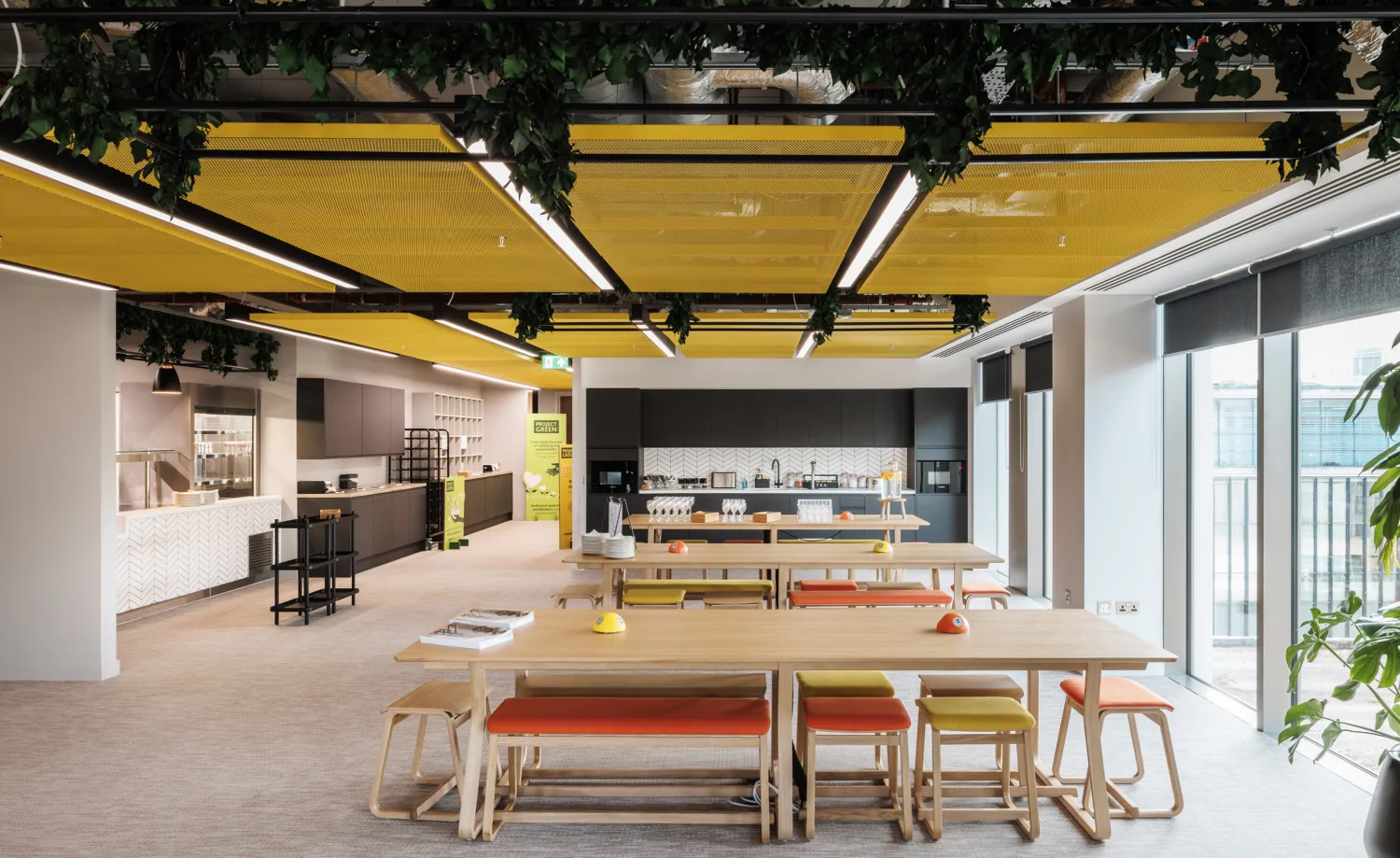 Staff eating and coffee area, yellow acoustic panelling ceiling with wooden tables with orange and yellow seating, as well as black and white units for the kitchen area