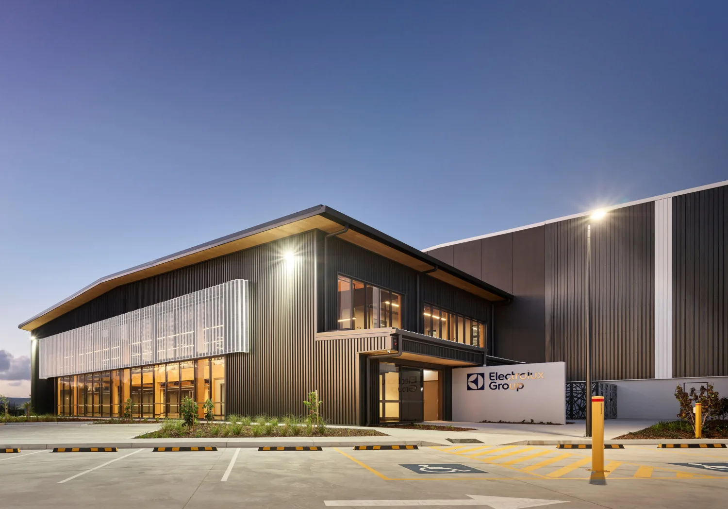 Exterior view of a modern industrial building with dark vertical cladding and large glass windows. The structure features an angled roof, illuminated façade, and landscaped area at the front. A sign near the entrance displays the text “Electrolux Group.” The foreground shows a paved driveway with marked parking bays and a pedestrian crossing.