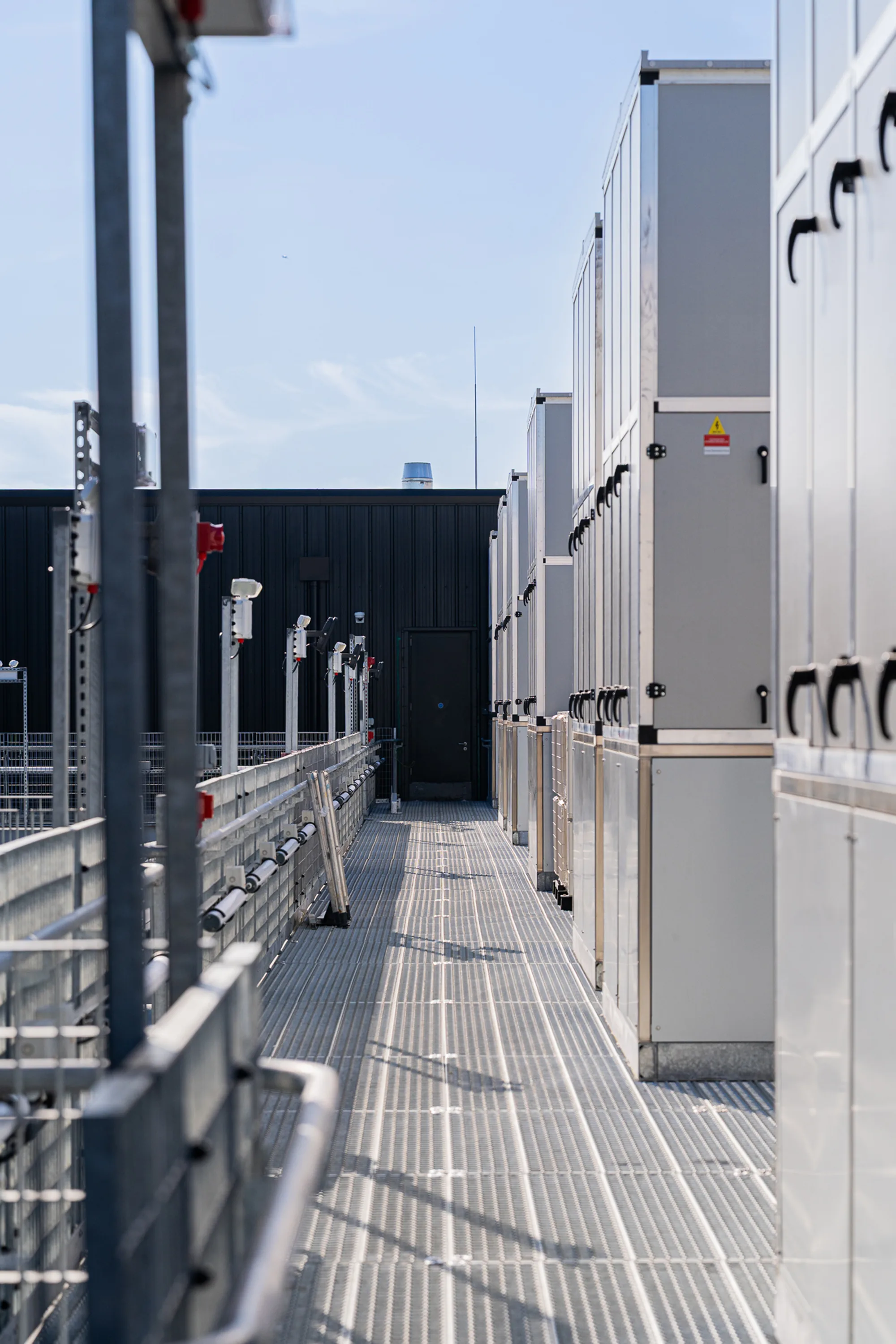 Narrow walkway with a metal grate floor between two rows of large grey industrial cabinets with black handles. Security cameras and lights are mounted on poles. A black wall with a closed door is in the background.