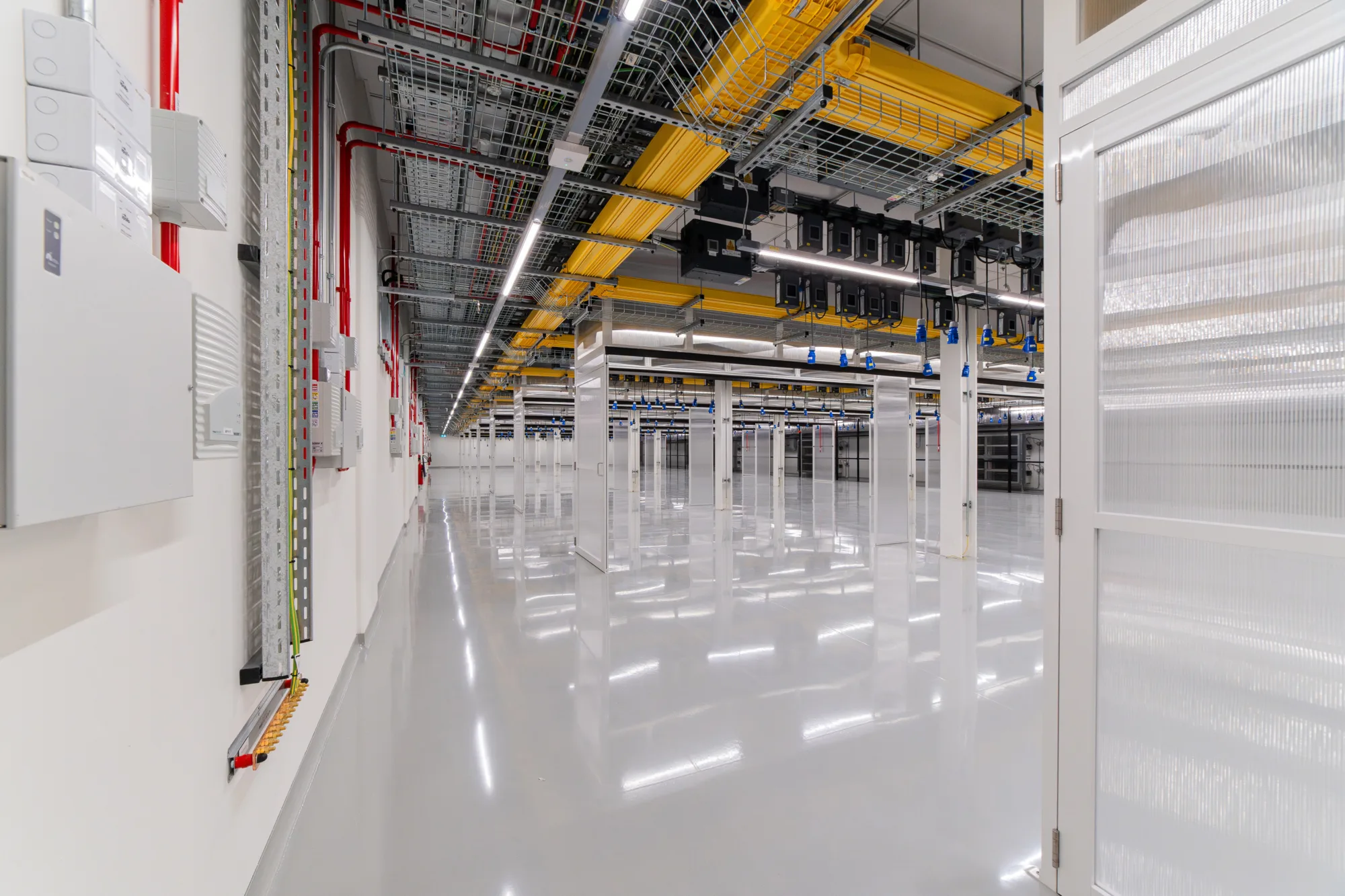 Spacious, brightly lit data centre with reflective floors, white server racks, yellow cable trays and electrical conduits on the ceiling, and red pipes and electrical panels on the left wall.