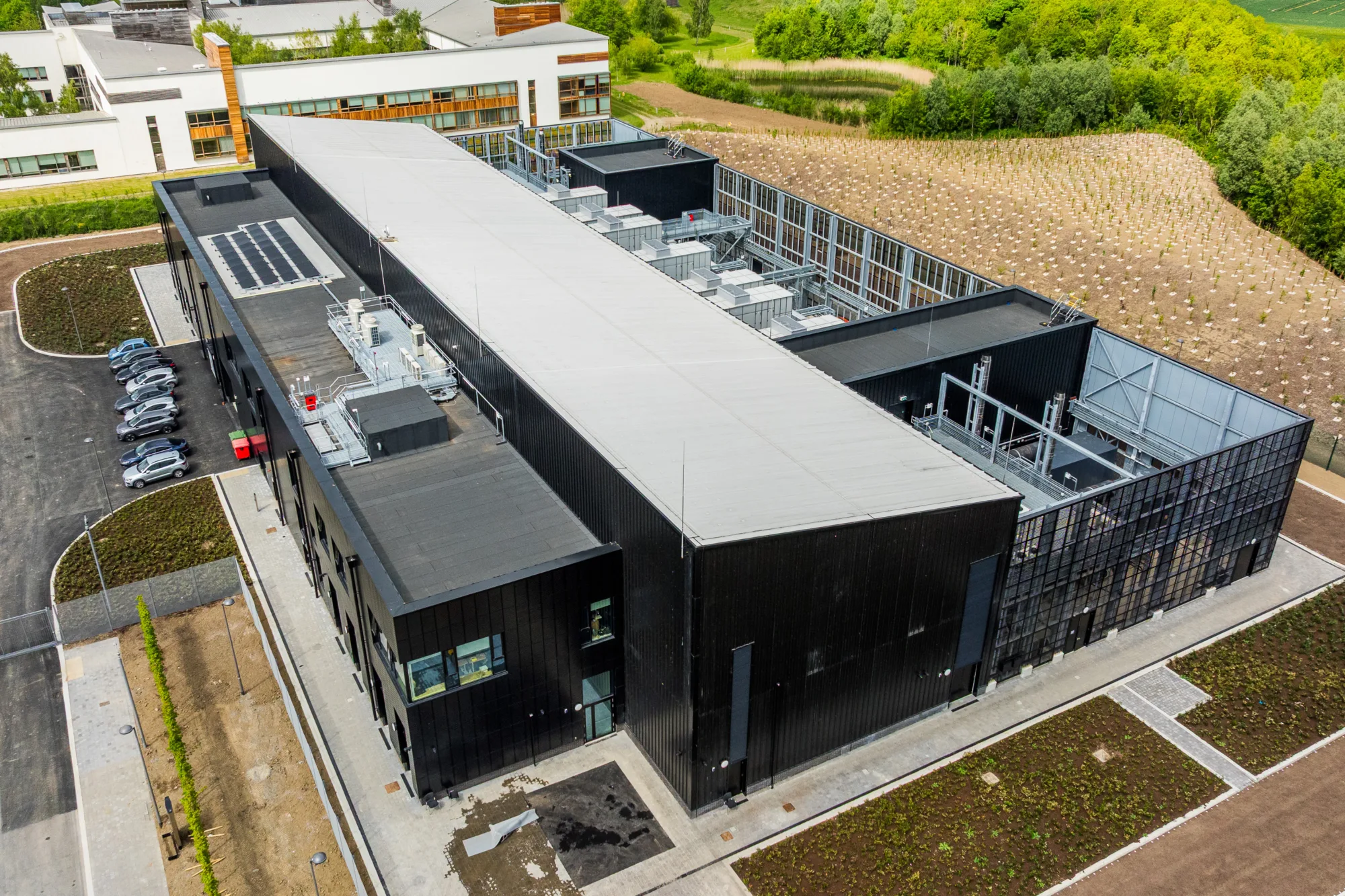 Aerial view of a large Data Centre building with a flat roof and black exterior, surrounded by paved areas, parked cars, and landscaped grounds. Green fields and a white building are visible in the background.